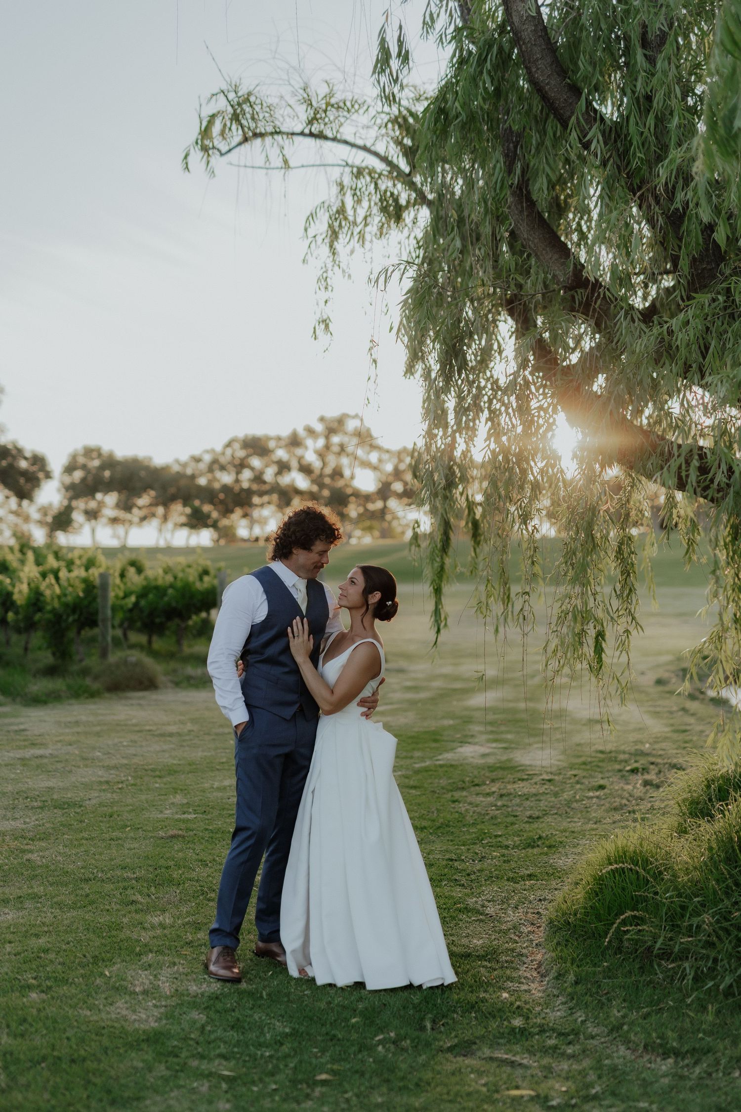 Bride and groom share romantic moment under weeping willow tree at golden hour vineyard wedding.