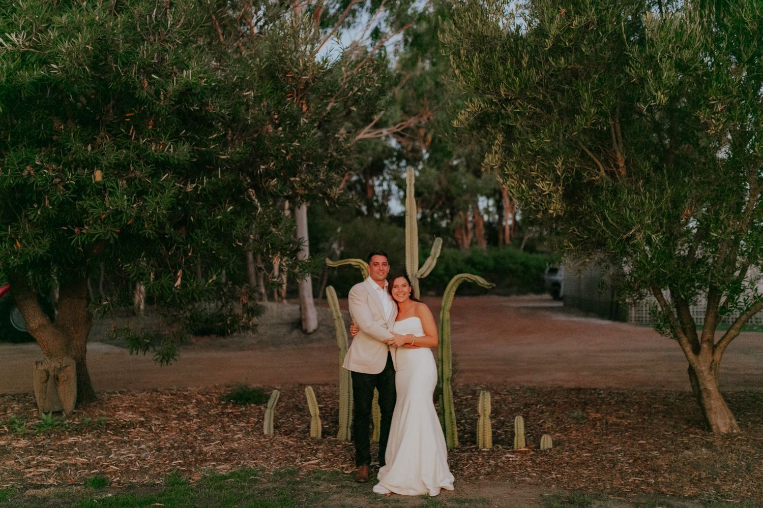Bride and groom embracing beside rustic wooden LOVE letters surrounded by eucalyptus trees at dusk.