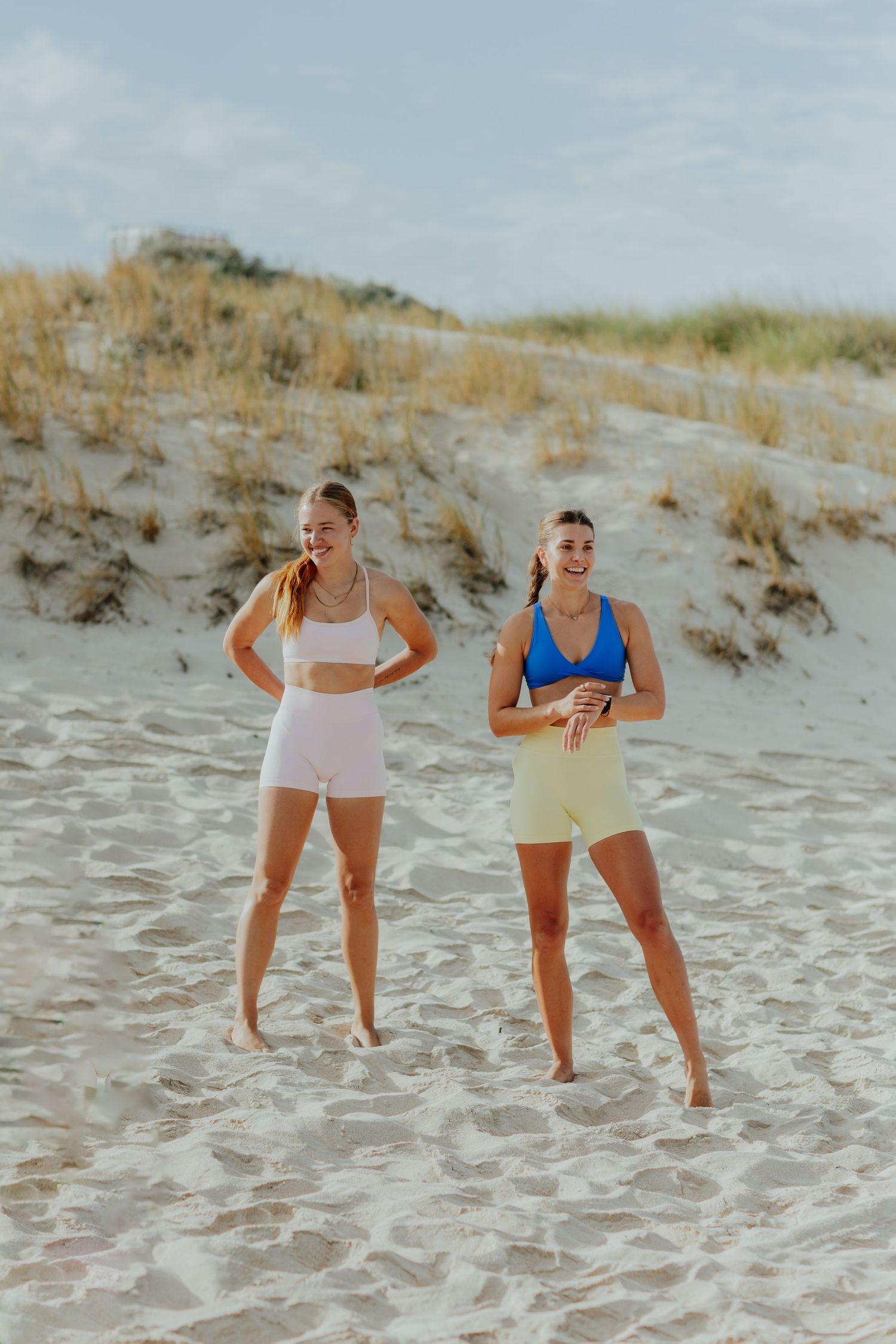 Two smiling women in colorful athletic wear work out together on a sandy beach with dunes.