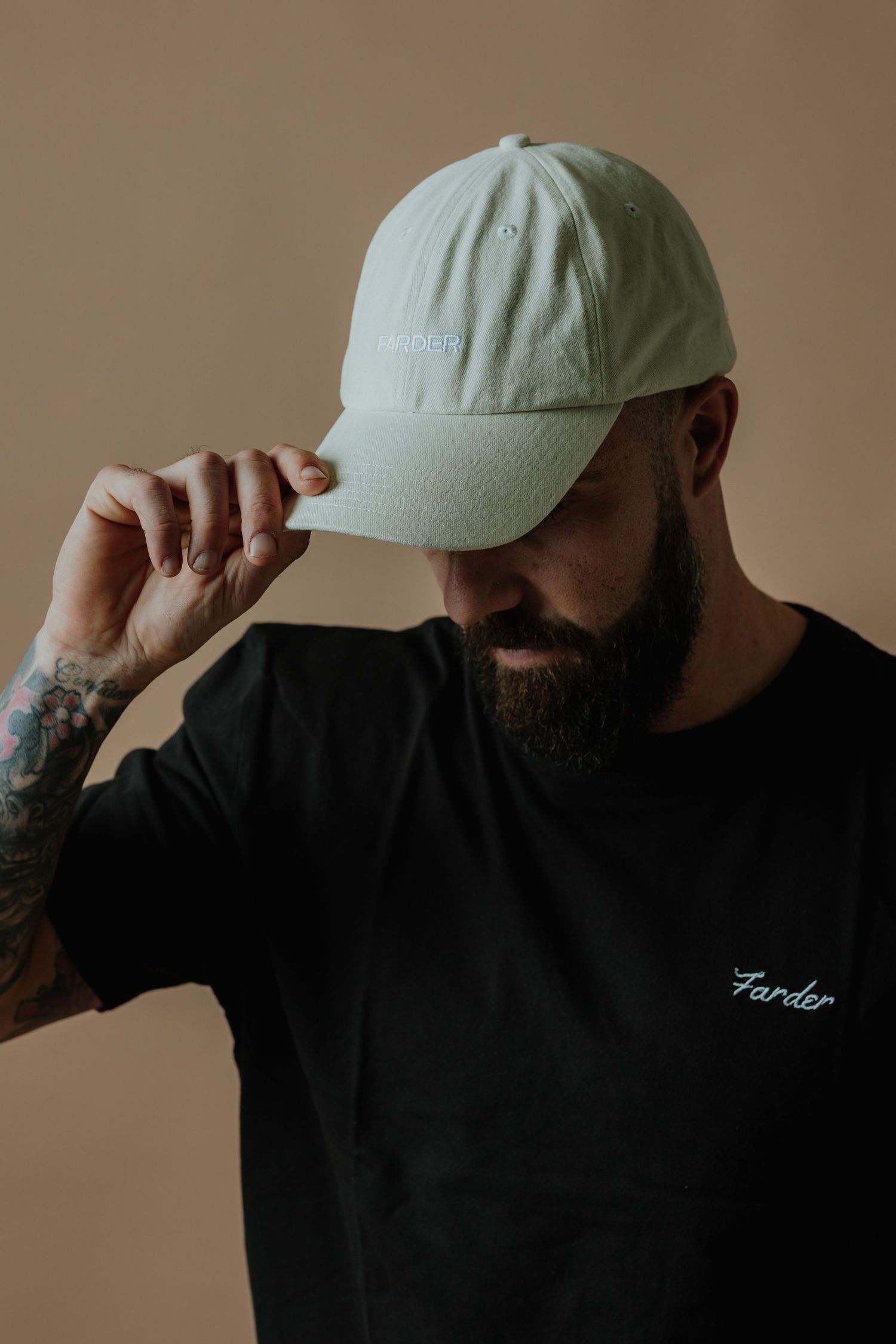 Tattooed bearded man adjusting a light green Farder branded baseball cap while wearing a black t-shirt.