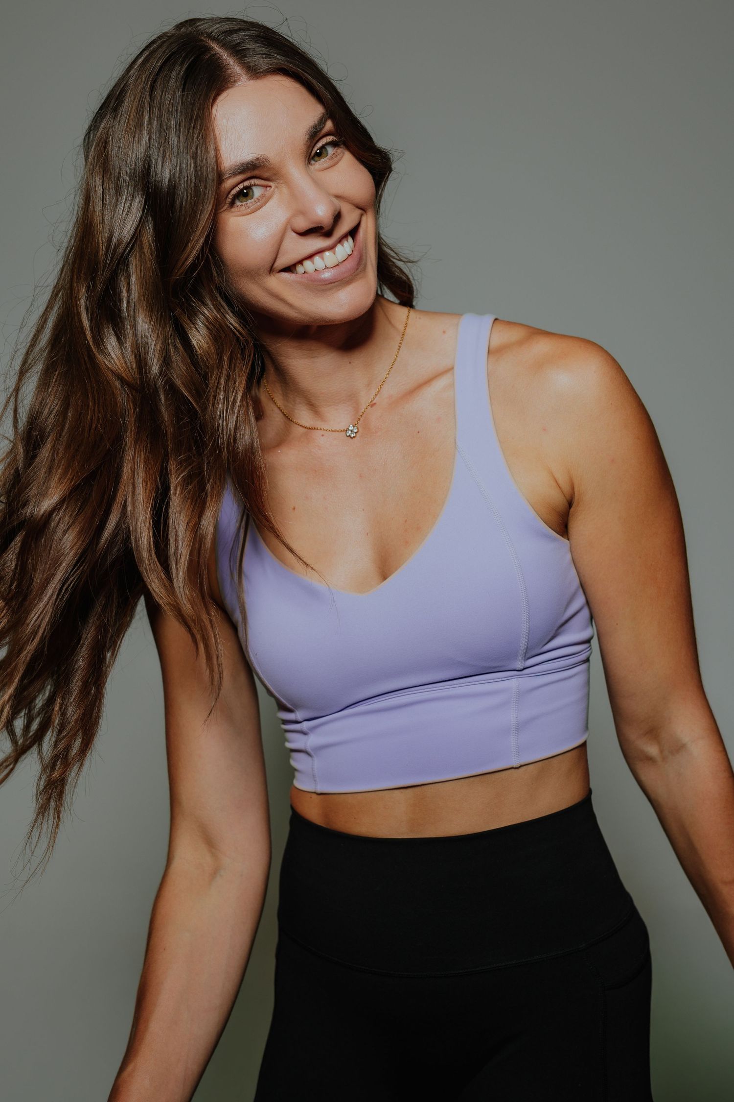 Smiling woman in lavender sports bra and black leggings posing against a gray background.