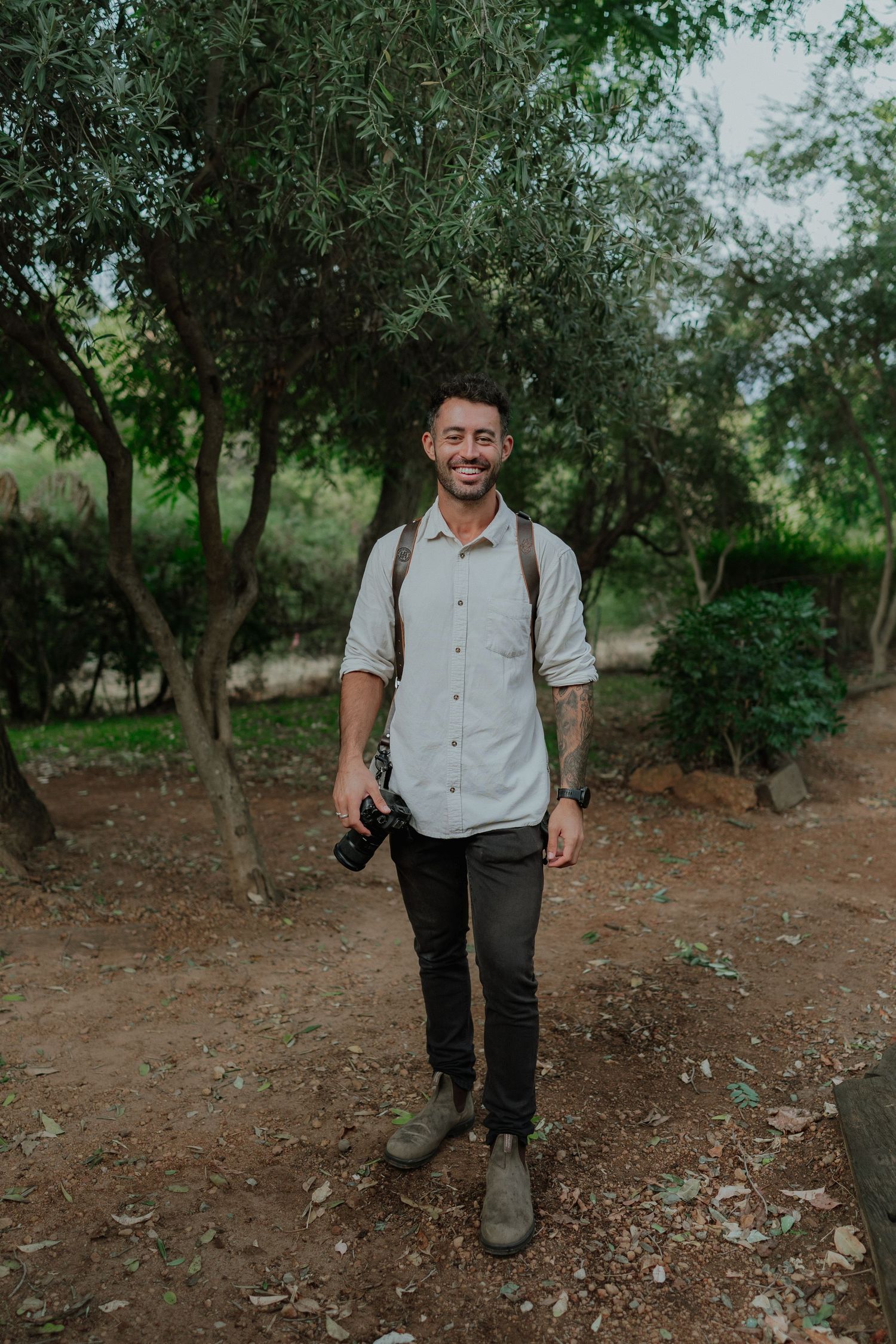 Smiling male photographer holding camera in lush green park wearing light shirt and backpack.