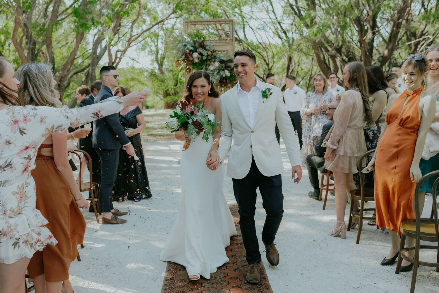 Newlyweds walking back down outdoor aisle as guests celebrate with confetti in bushland setting.