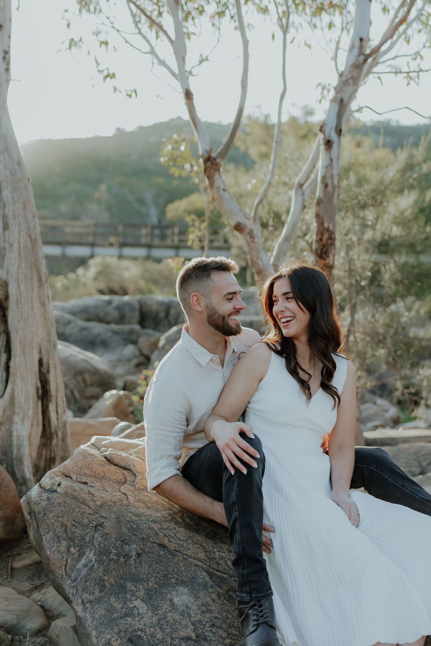 Happy couple sitting on rocks laughing together in golden sunlight with trees and bridge behind them.