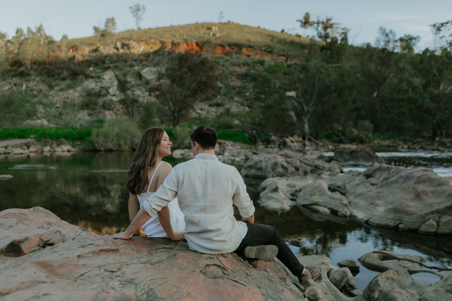 Romantic couple sitting together on rocks overlooking a peaceful river with a scenic landscape.
