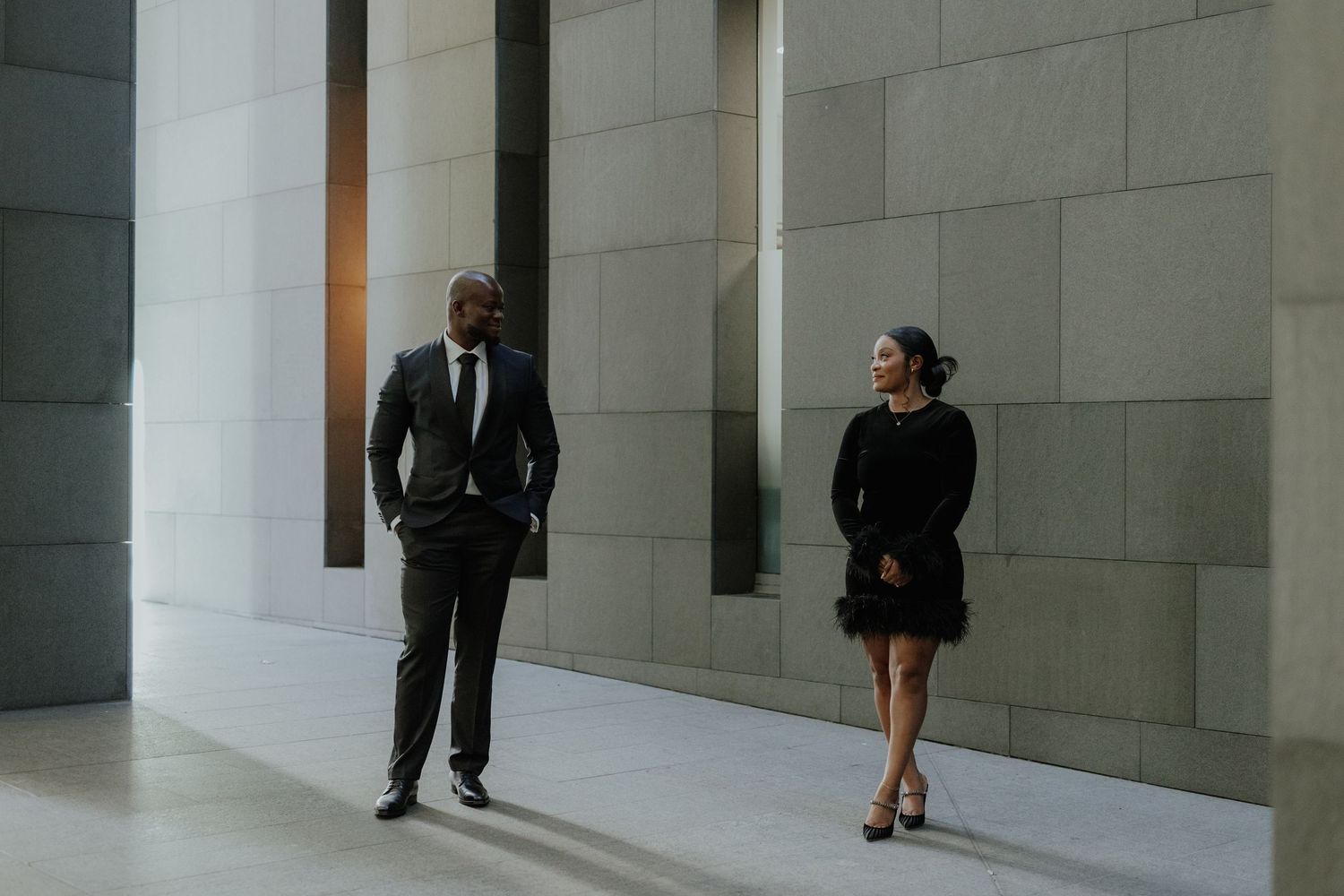 Elegant interracial couple in black formal wear walking in modern architectural space.