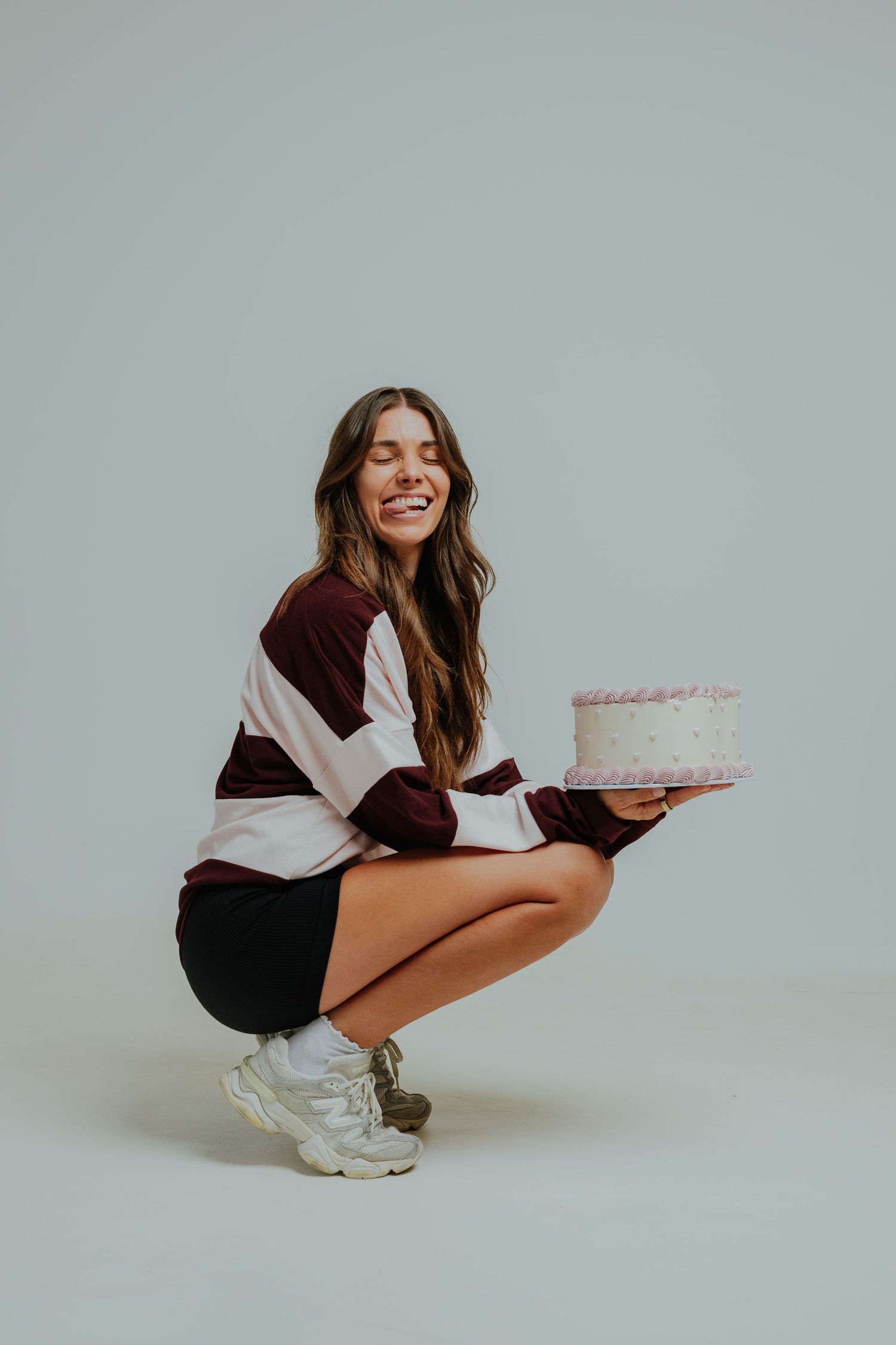 Smiling woman crouching and holding a pink birthday cake on a light gray background.