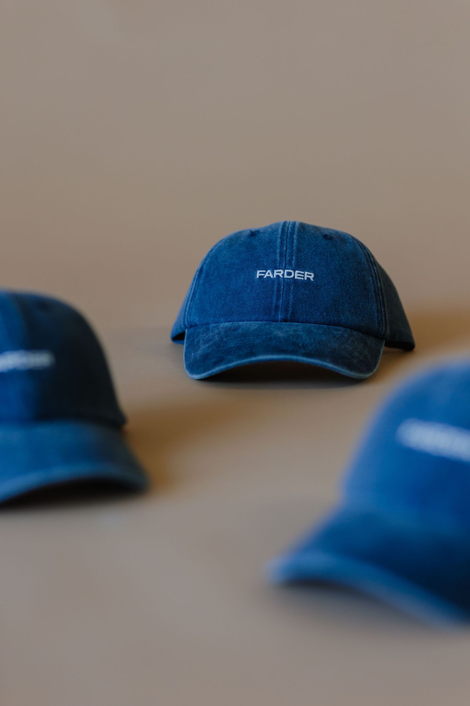 Three blue denim Farder branded baseball caps arranged on a neutral beige surface.