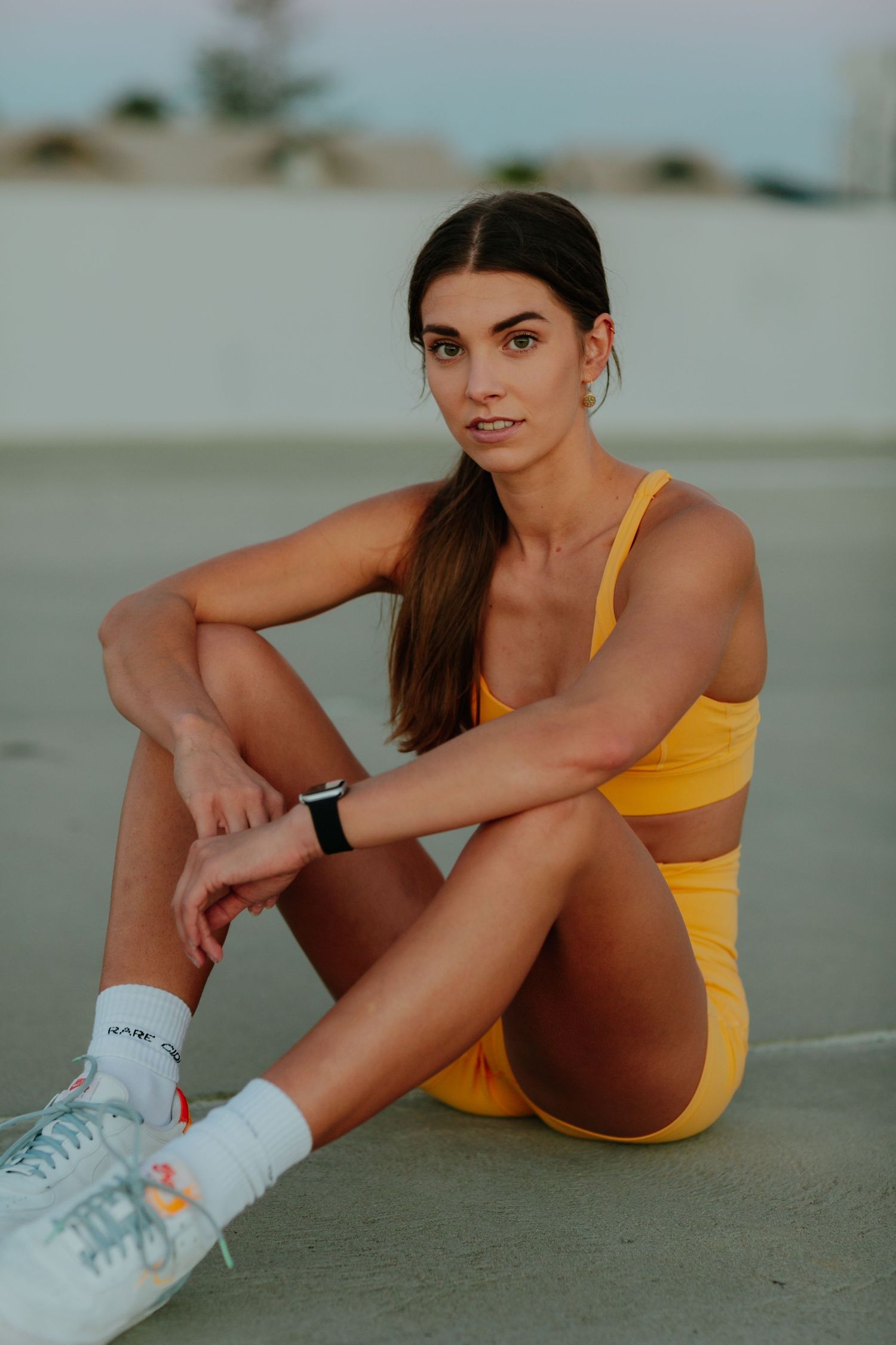 Fit woman in yellow activewear sits relaxed on pavement wearing white sneakers and a smartwatch.