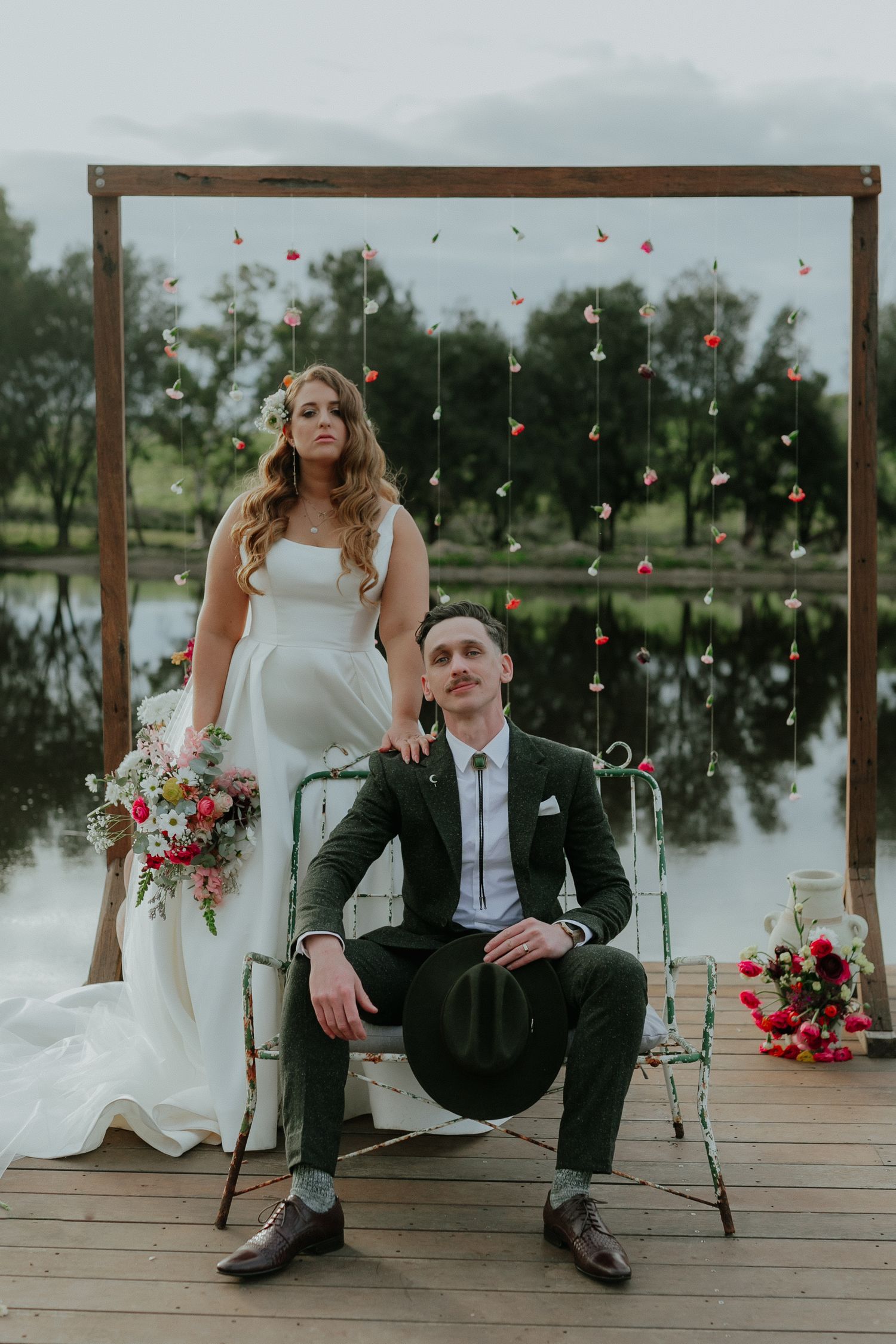 Bride and groom pose elegantly by lakeside floral arch backdrop at romantic outdoor wedding.