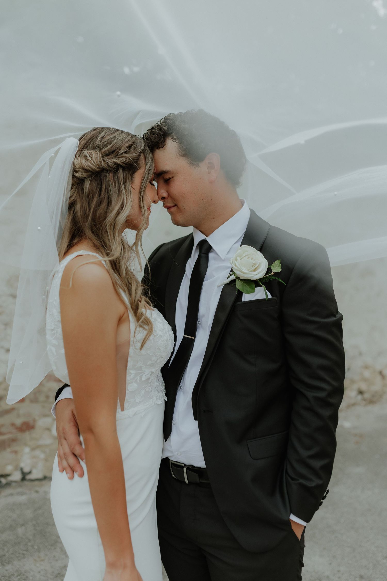 Bride and groom sharing an intimate forehead touch under a flowing veil outdoors.