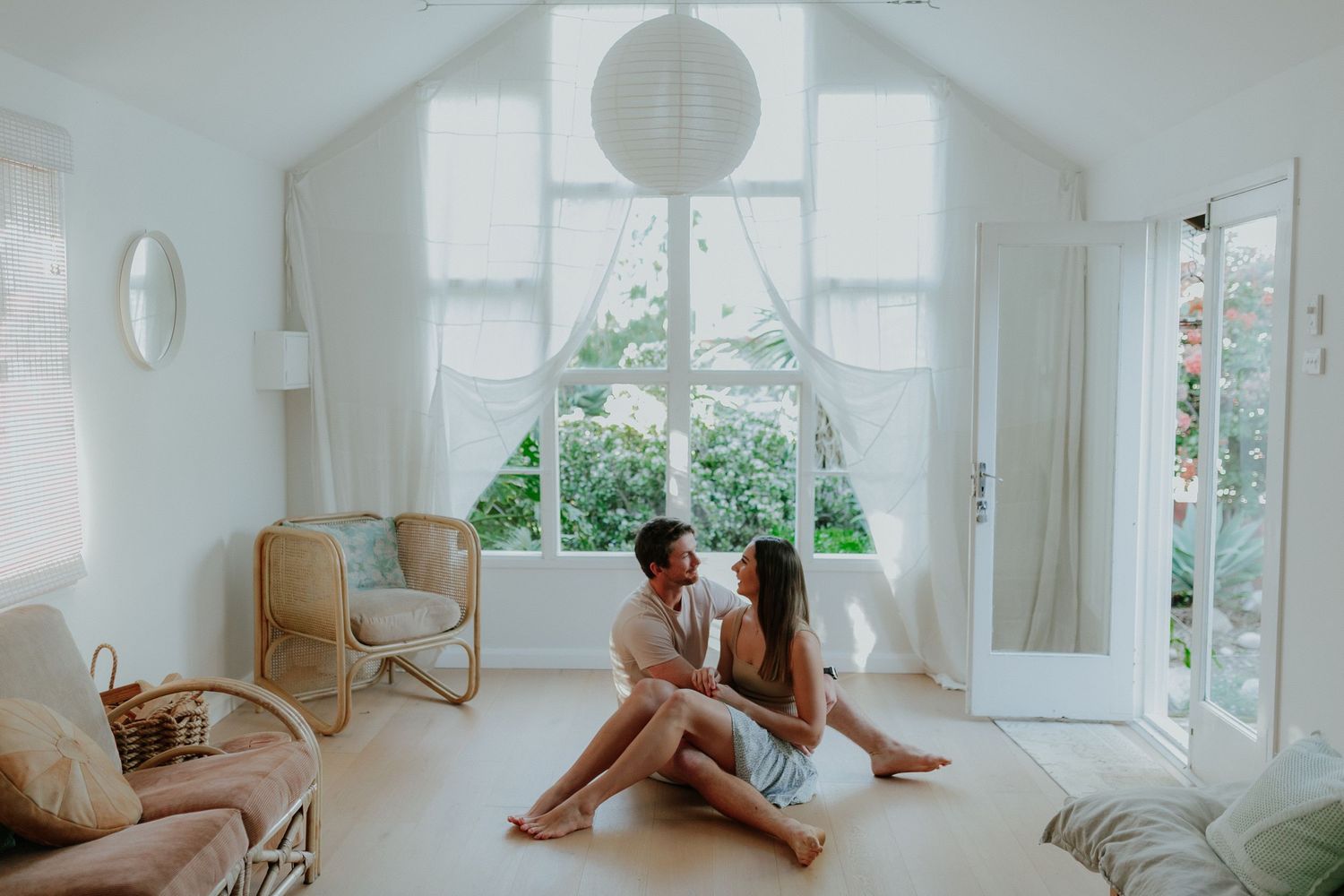 Happy couple sitting together on floor of bright white boho living room, sharing an intimate moment indoors.