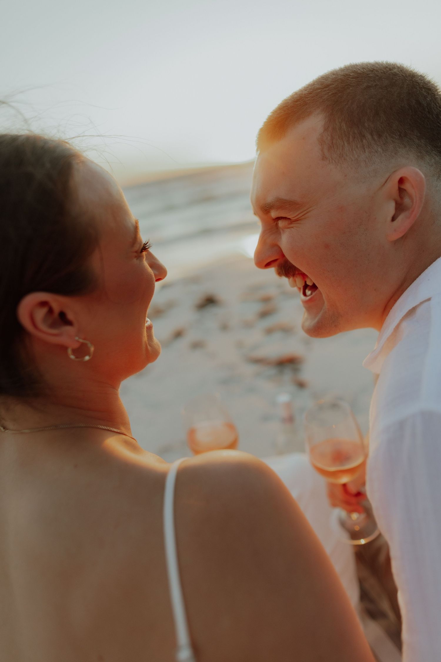 Laughing couple holding wine glasses facing each other on a beach at sunset.