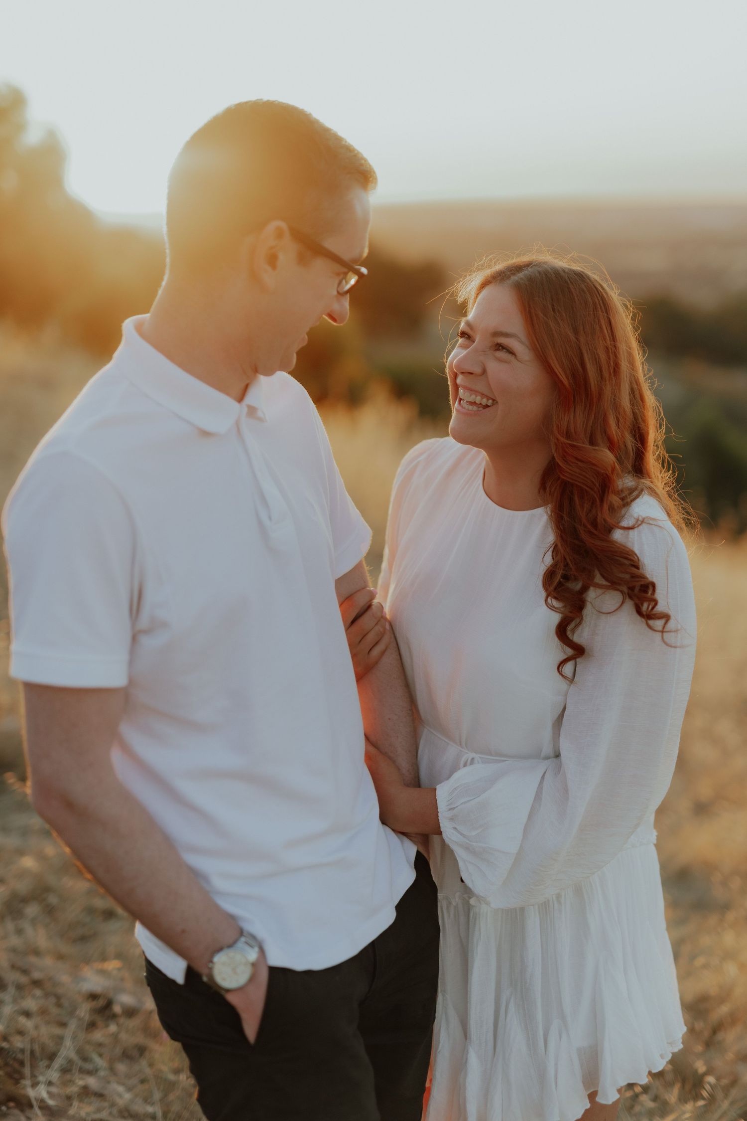 Smiling couple in white outfits embracing in a golden sunset field.