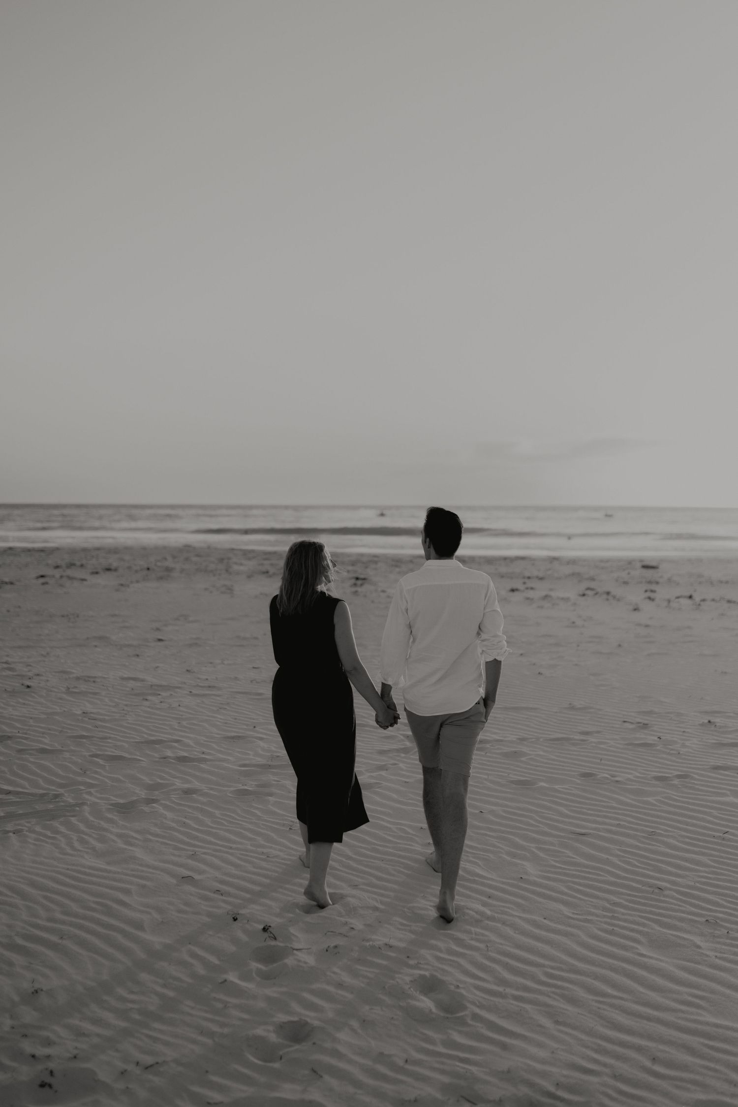 Black and white photo of couple holding hands walking toward ocean on wide sandy beach at dusk.