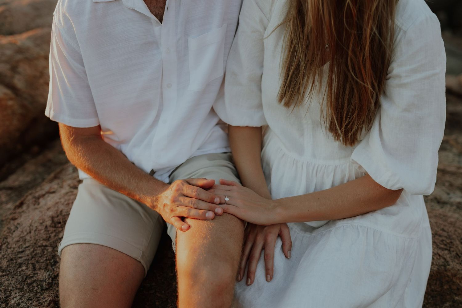 Close-up of couple sitting together showing engagement ring on woman's hand against white outfits.