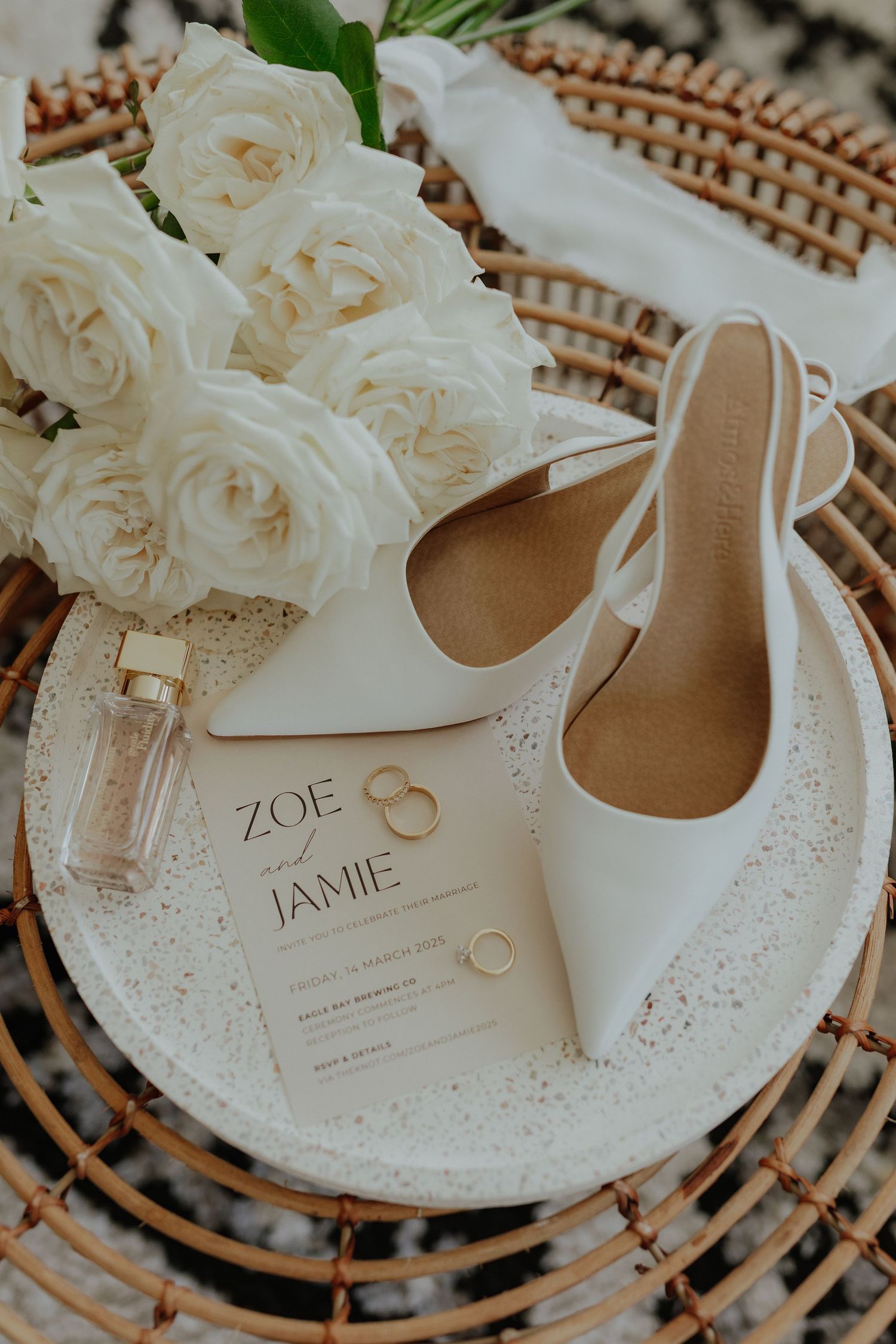 Flat lay of white bridal heels, wedding invitation for Zoe and Jamie, rings, and roses.