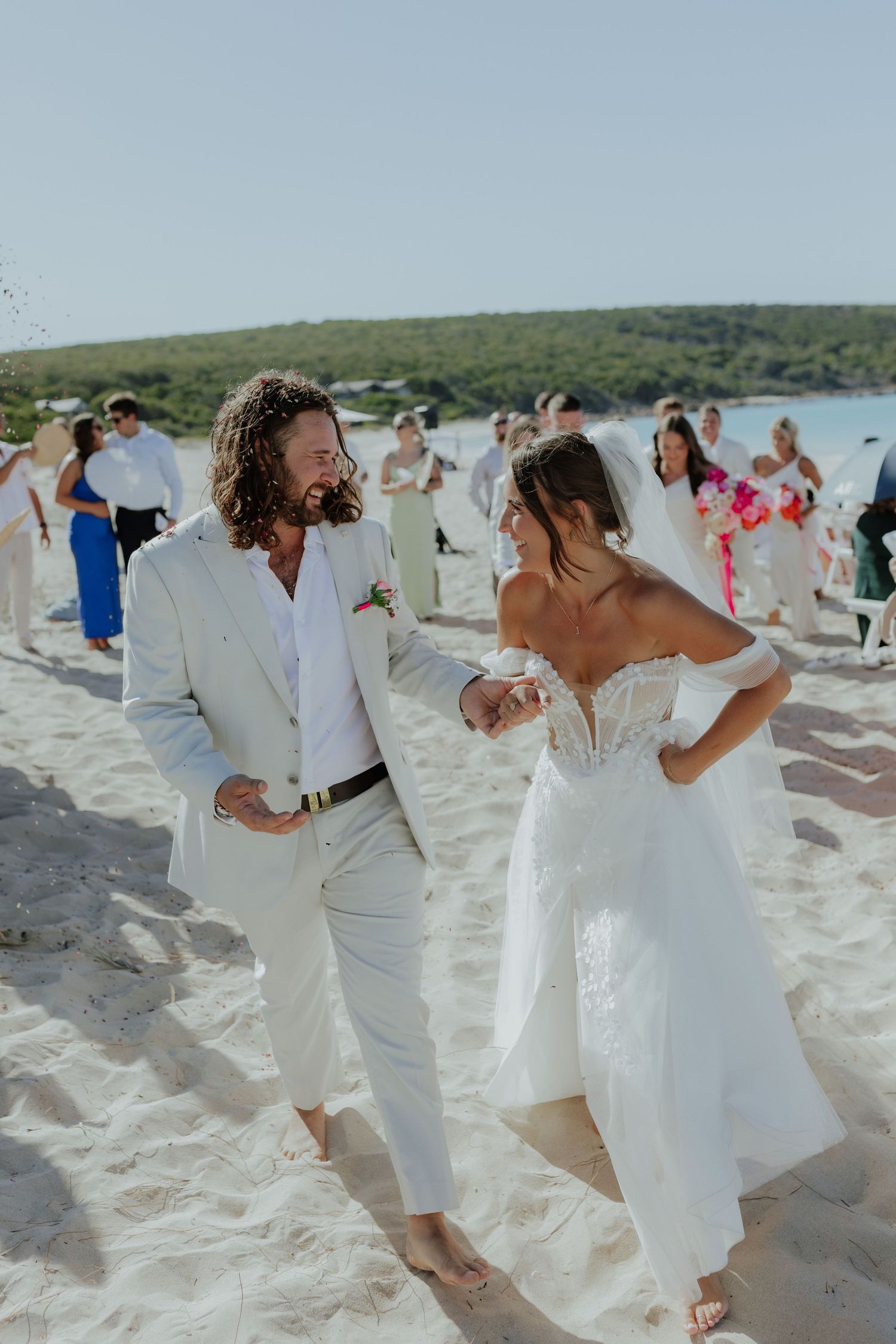 Bride and groom walking barefoot on white sand beach, smiling joyfully in elegant white wedding attire.