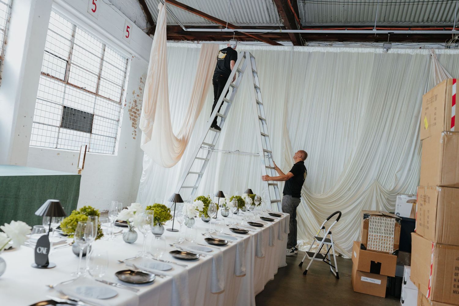 Event setup in progress with a worker on a ladder hanging white drapes behind a formally set dining table.