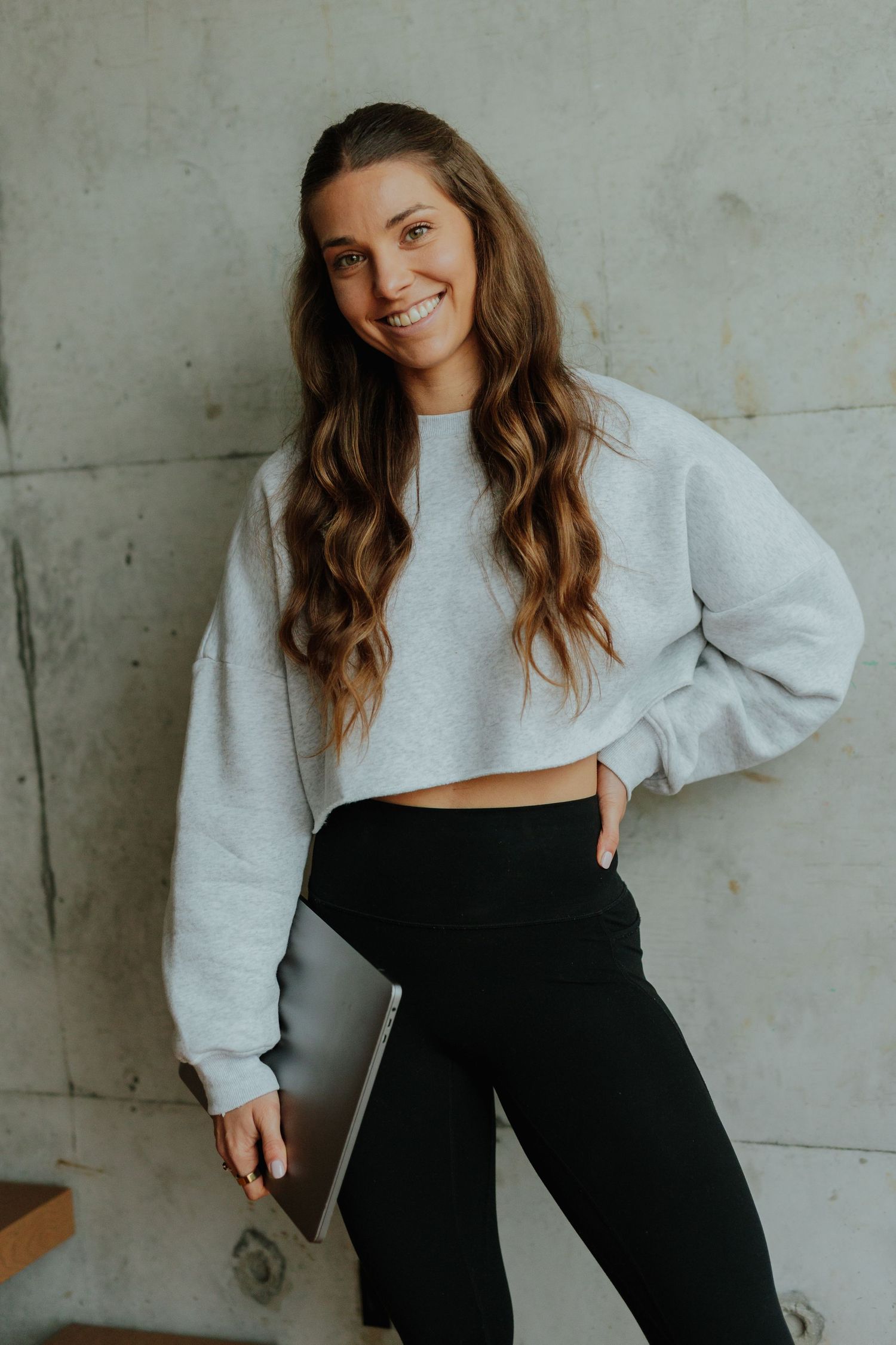 Happy young woman in grey cropped sweatshirt holding laptop while leaning against concrete wall.