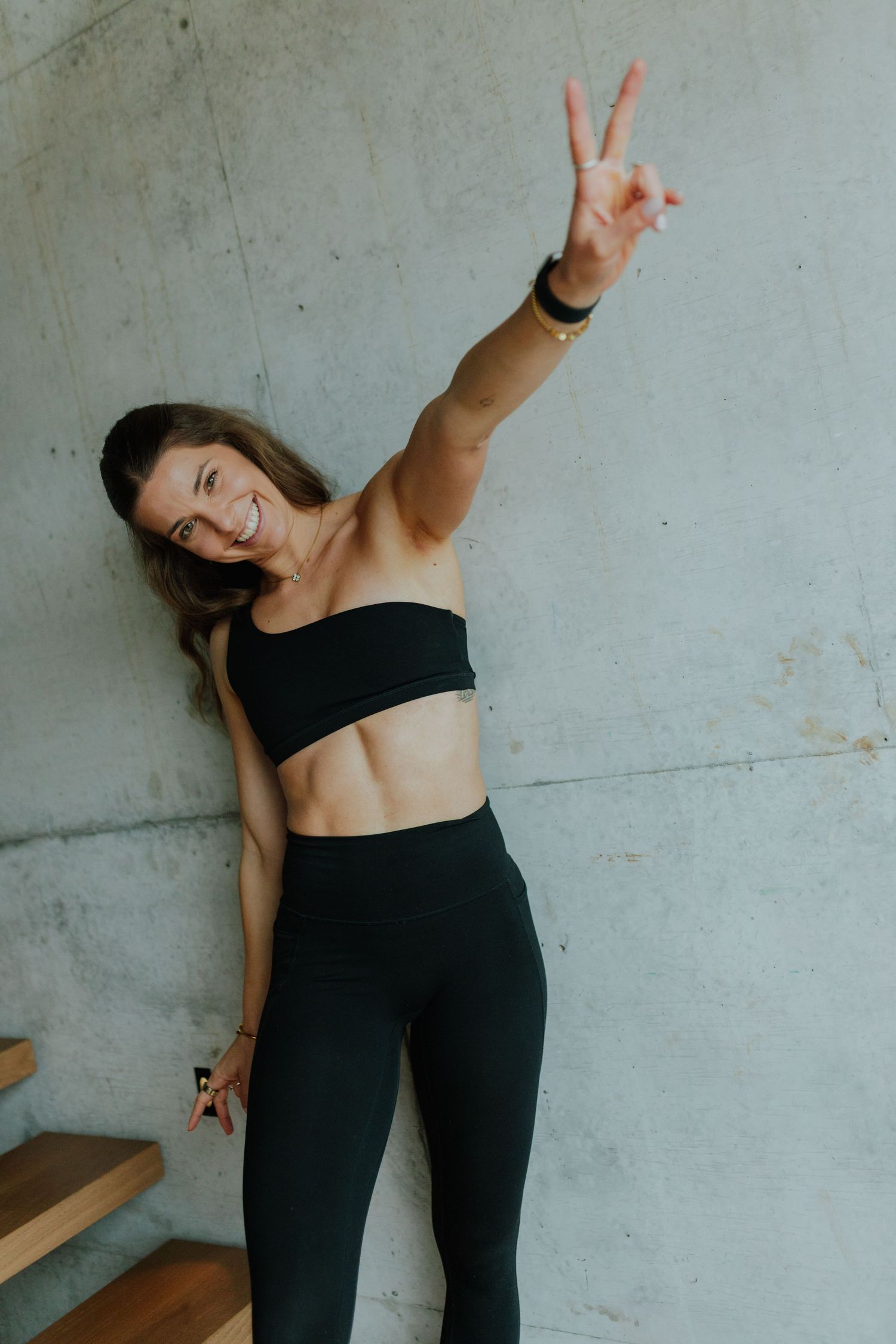 Fit woman in black bandeau top and leggings making peace sign against concrete wall.