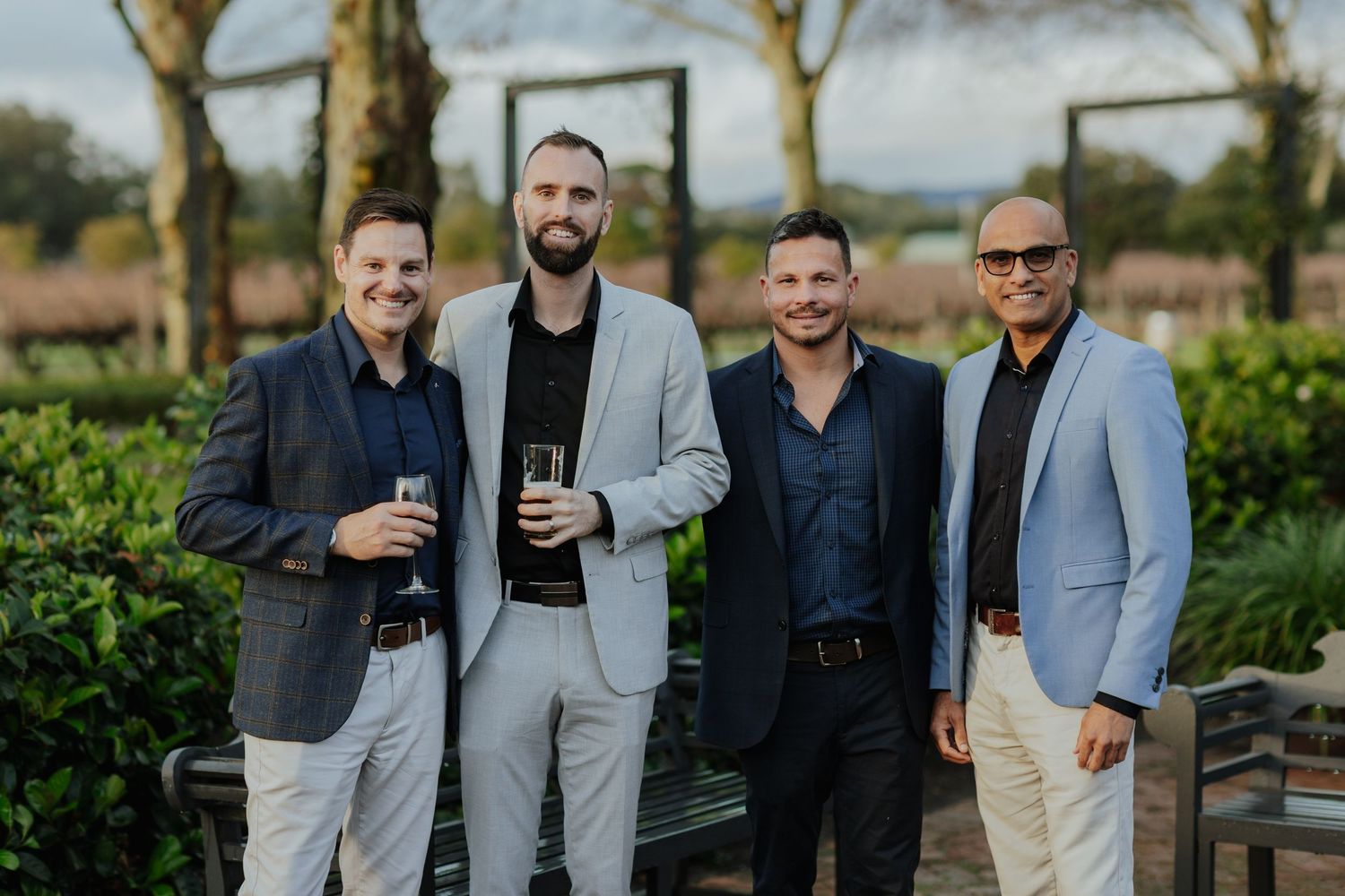 Four men in smart casual blazers smiling together outdoors at a vineyard event.