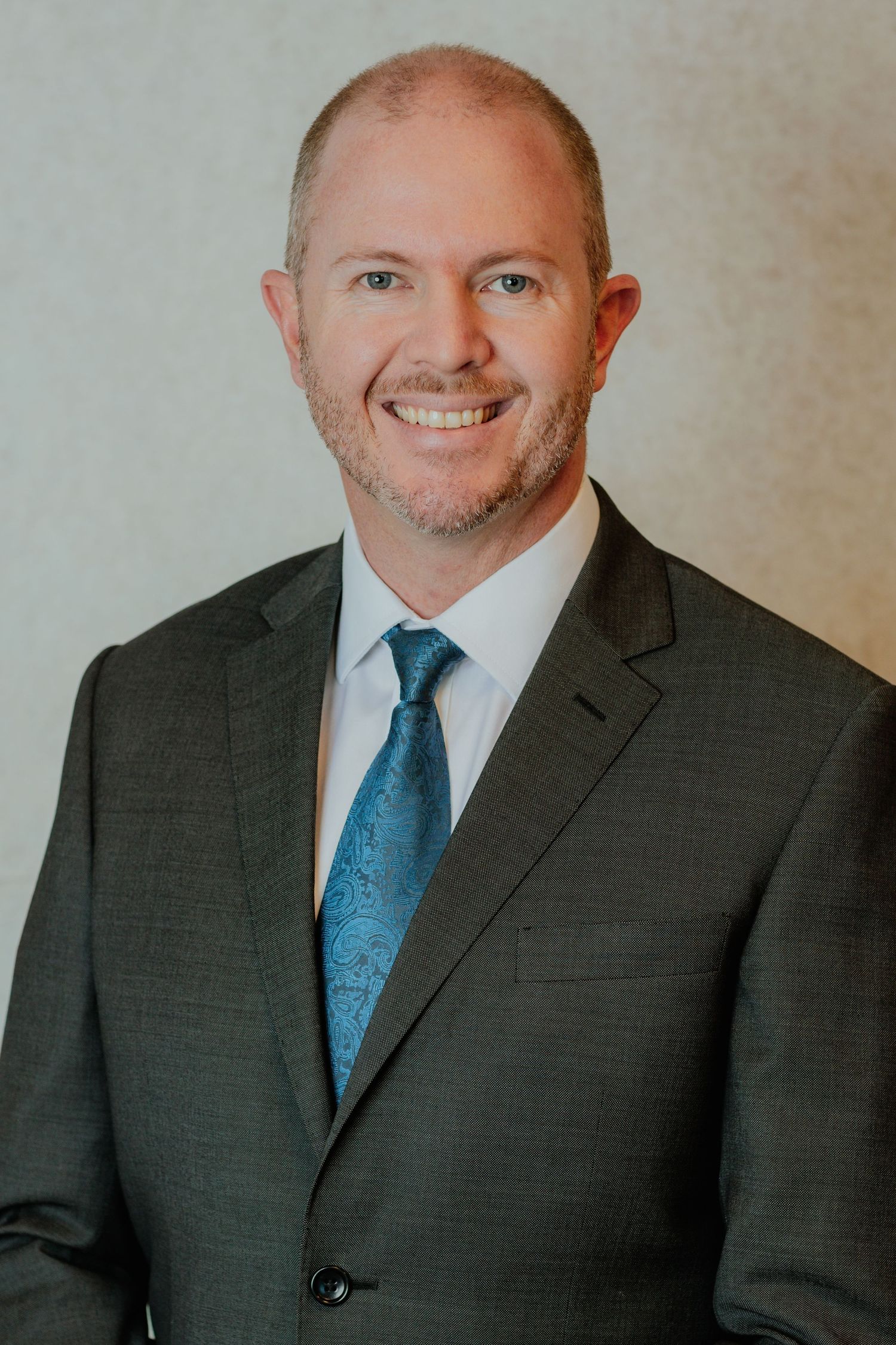 Smiling professional businessman in grey suit and teal tie poses for a corporate headshot.