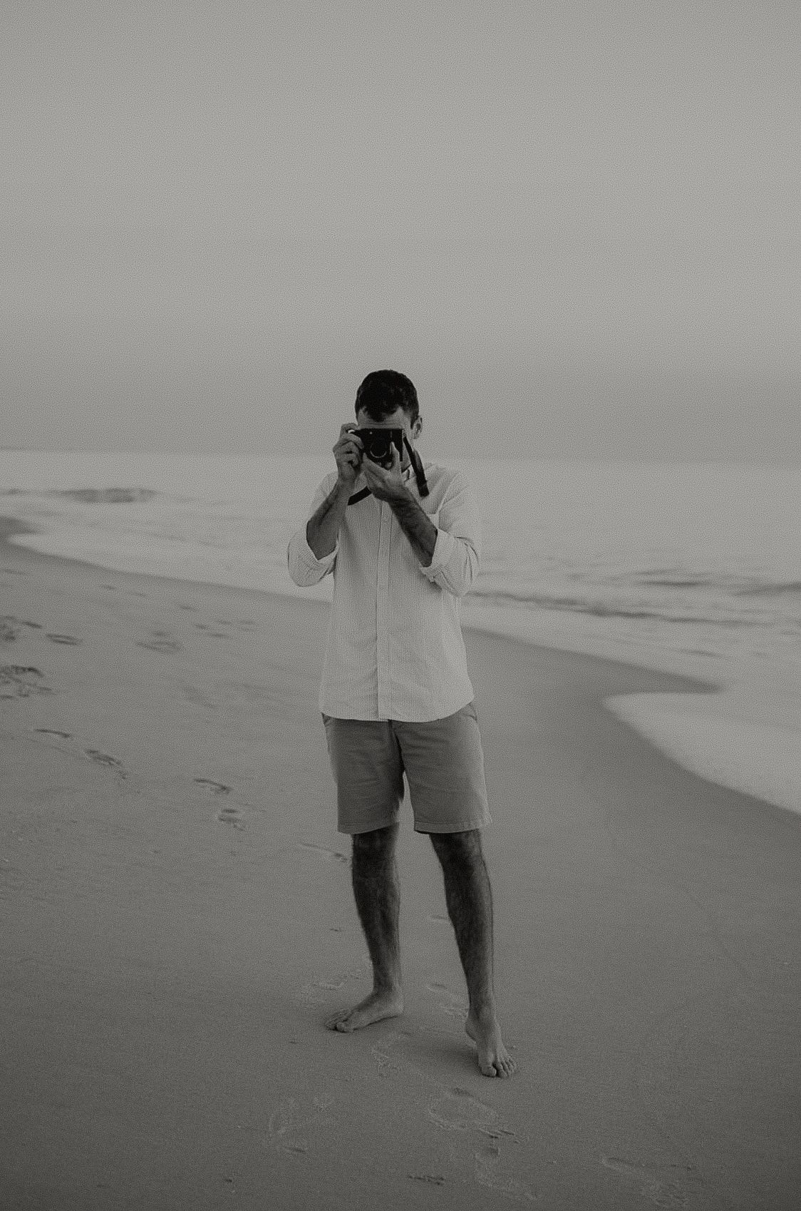 Man standing barefoot on misty beach holding camera up to face in black and white.