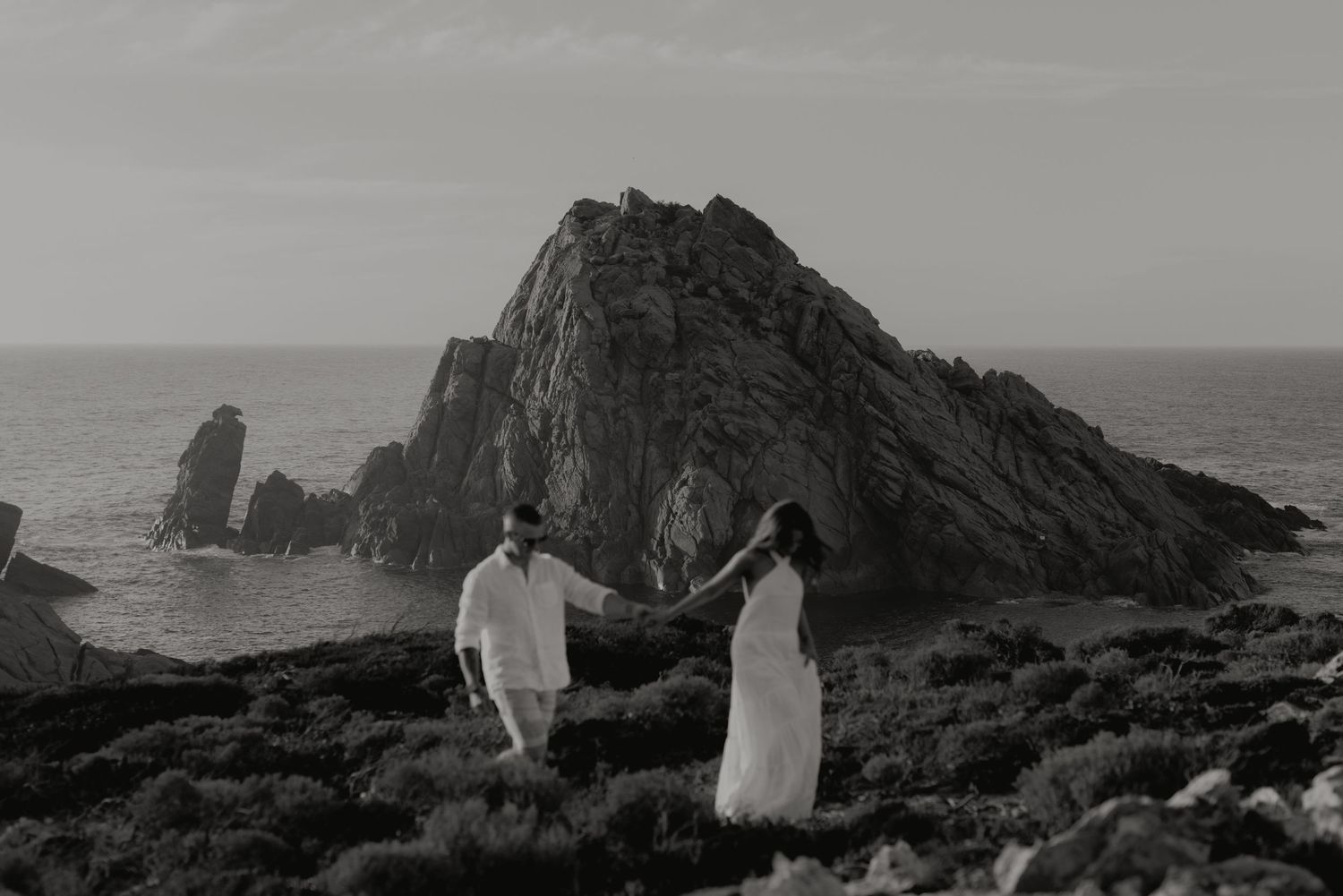 Black and white photo of couple holding hands near dramatic coastal rock formation by the sea.