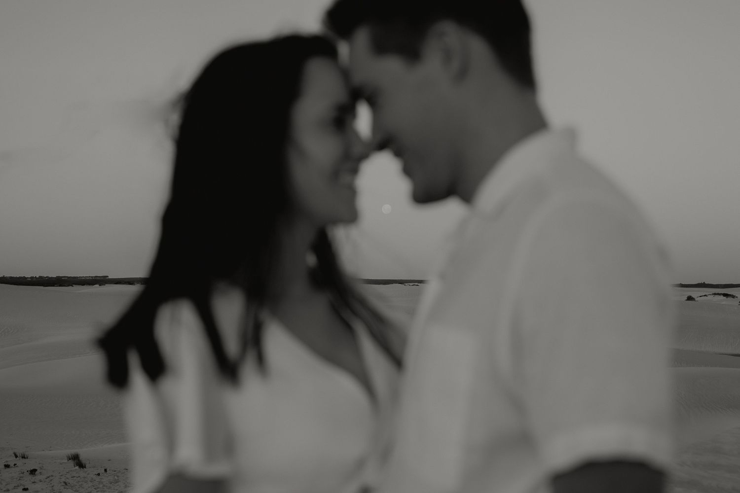 Soft blurred black and white photo of a couple touching foreheads intimately near a beach shoreline.