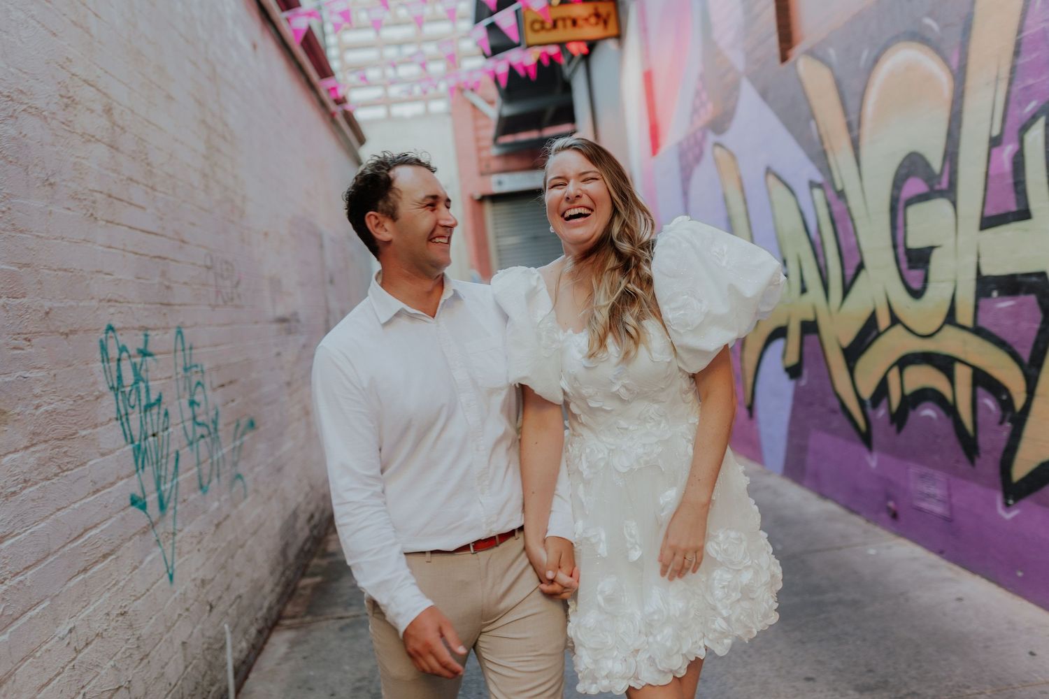 Laughing couple holding hands walking through a colorful graffiti alley, woman in white ruffled mini dress.