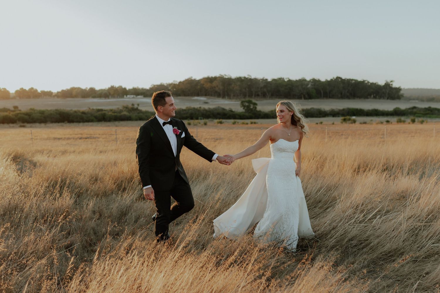 Bride and groom holding hands walking through golden wheat fields at sunset together.
