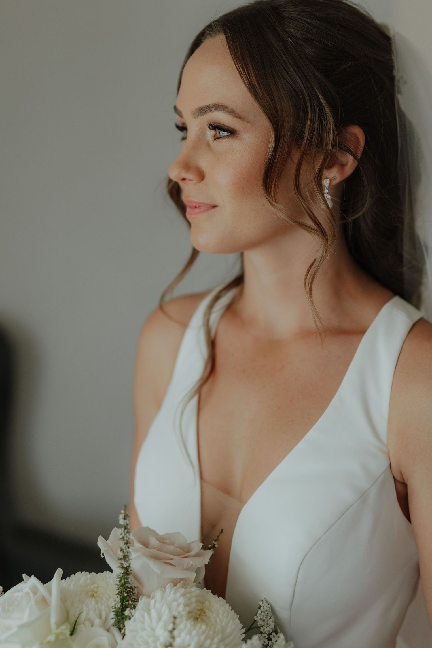 Elegant bride in sleek V-neck white gown holding white floral bouquet, gazing softly to the side.
