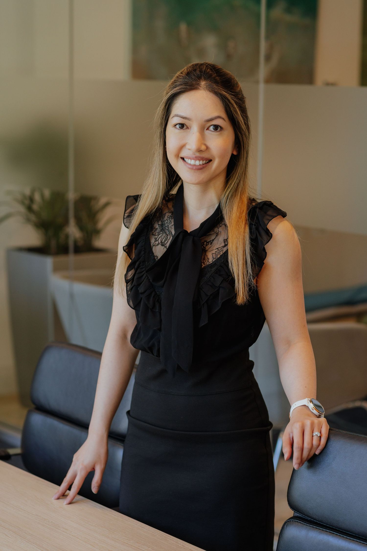 Smiling professional woman in black ruffled top leaning on a conference room table.