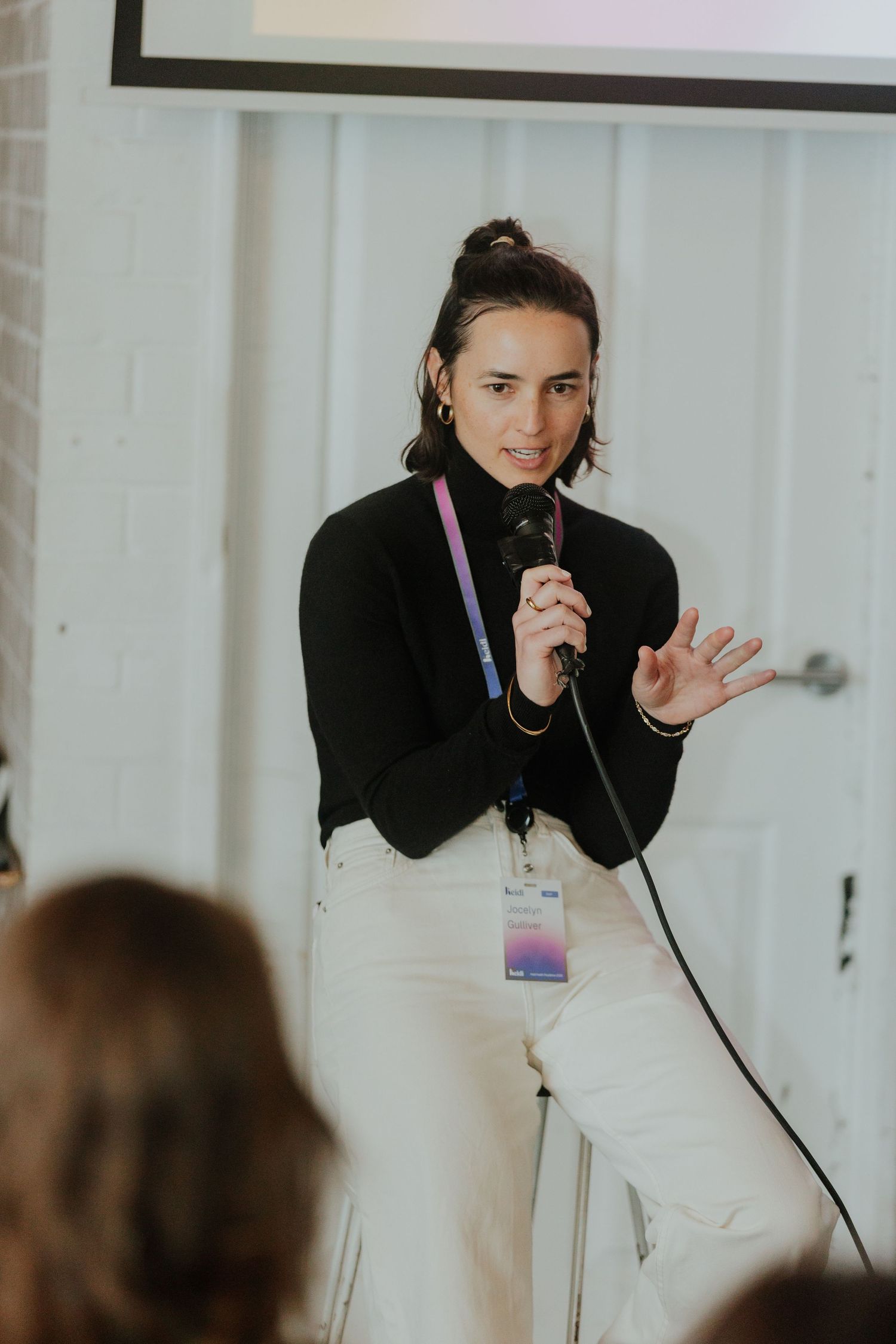 Young woman in black turtleneck speaking into microphone at a conference with lanyard badge.