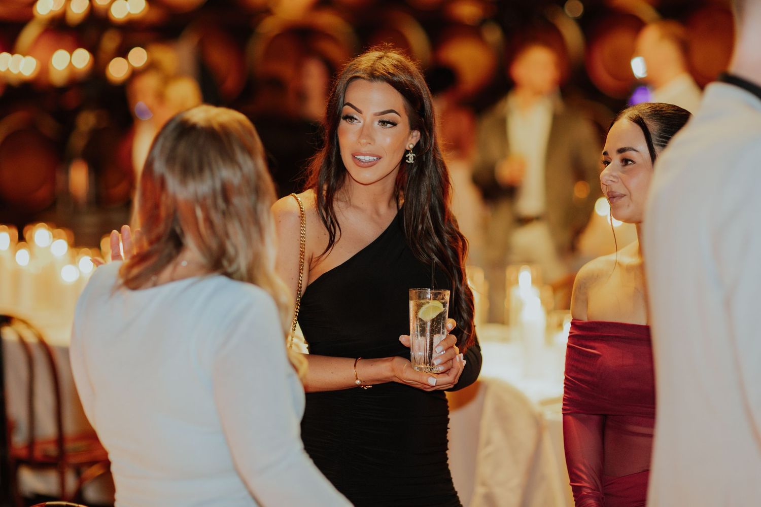 Elegant woman in black one-shoulder dress holding champagne at a candlelit winery event.