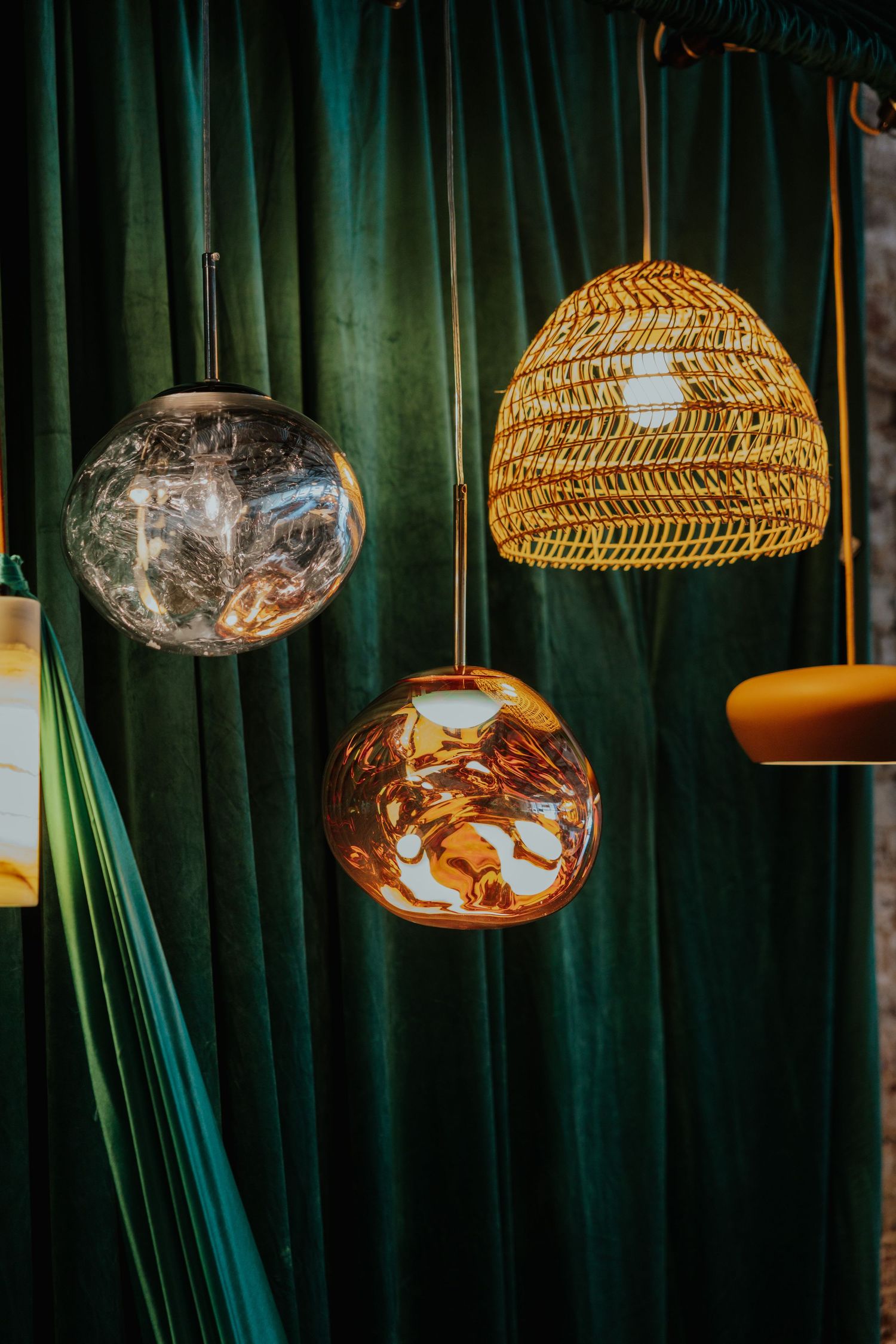 Three decorative pendant lights including wicker and amber glass globe styles displayed against green velvet curtains.