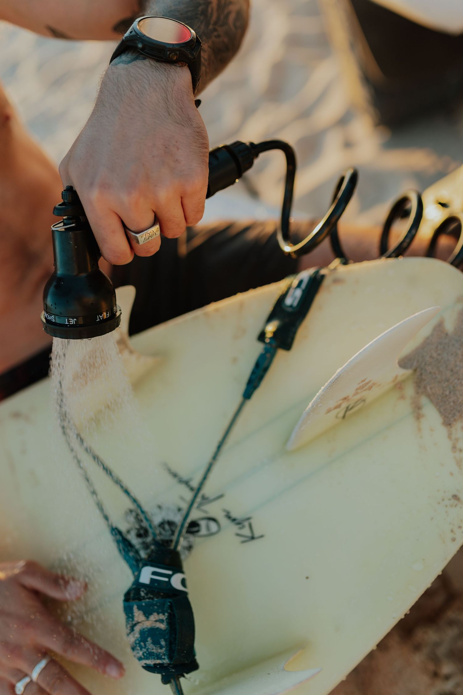 Close-up of tattooed hand attaching a leash to a surfboard on sandy beach.