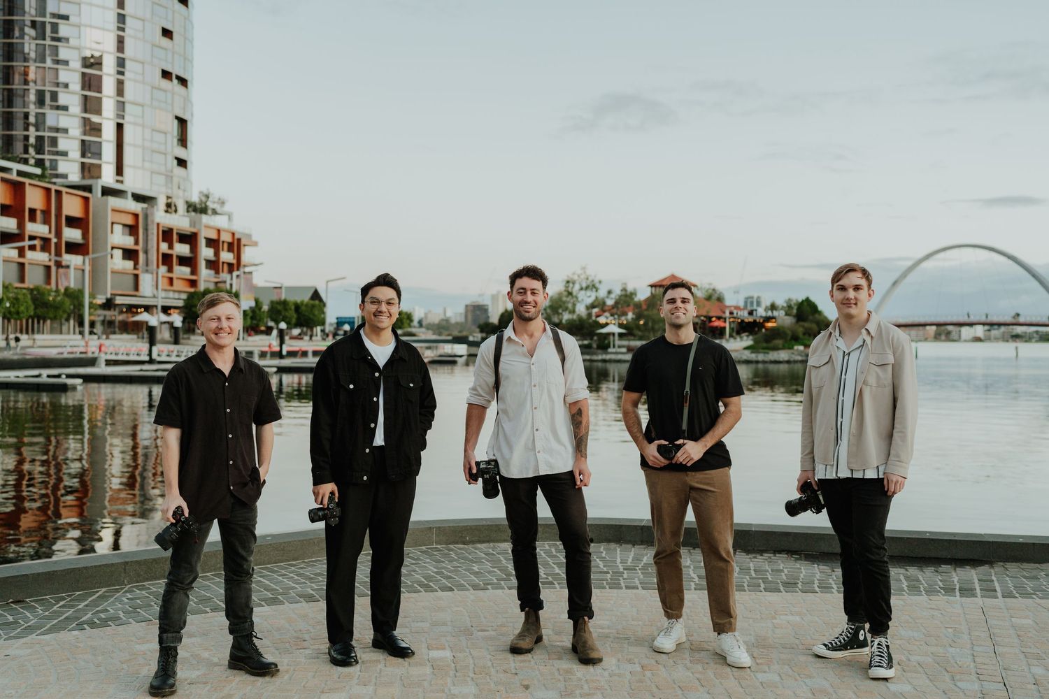 Group of five photographers posing by riverside with city buildings at dusk in Perth, Australia.