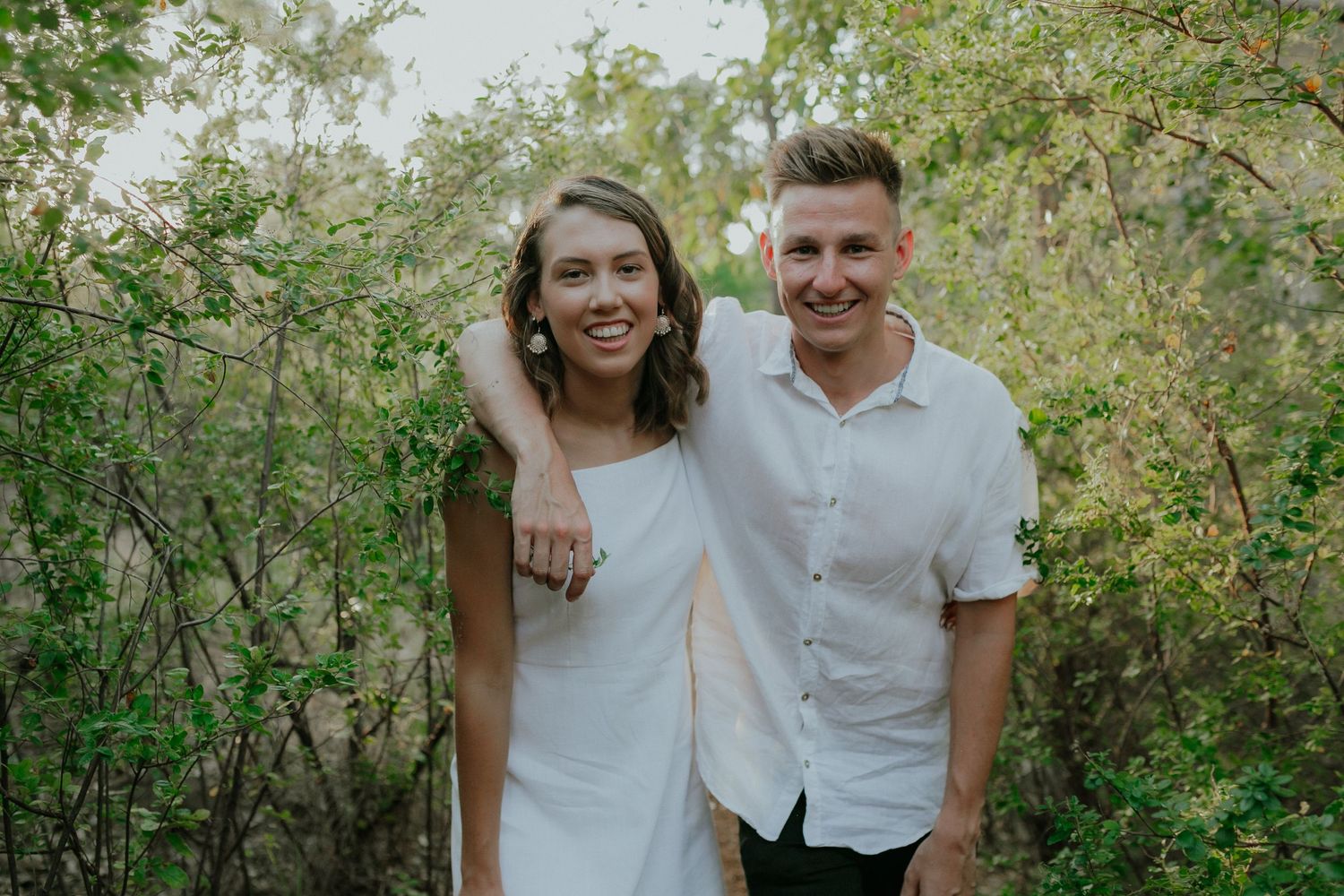 Smiling couple posing together in a natural green outdoor setting for an engagement photo.