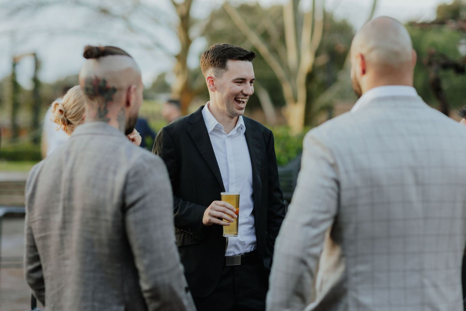 Young man in black suit laughing while socializing with guests at an outdoor event.