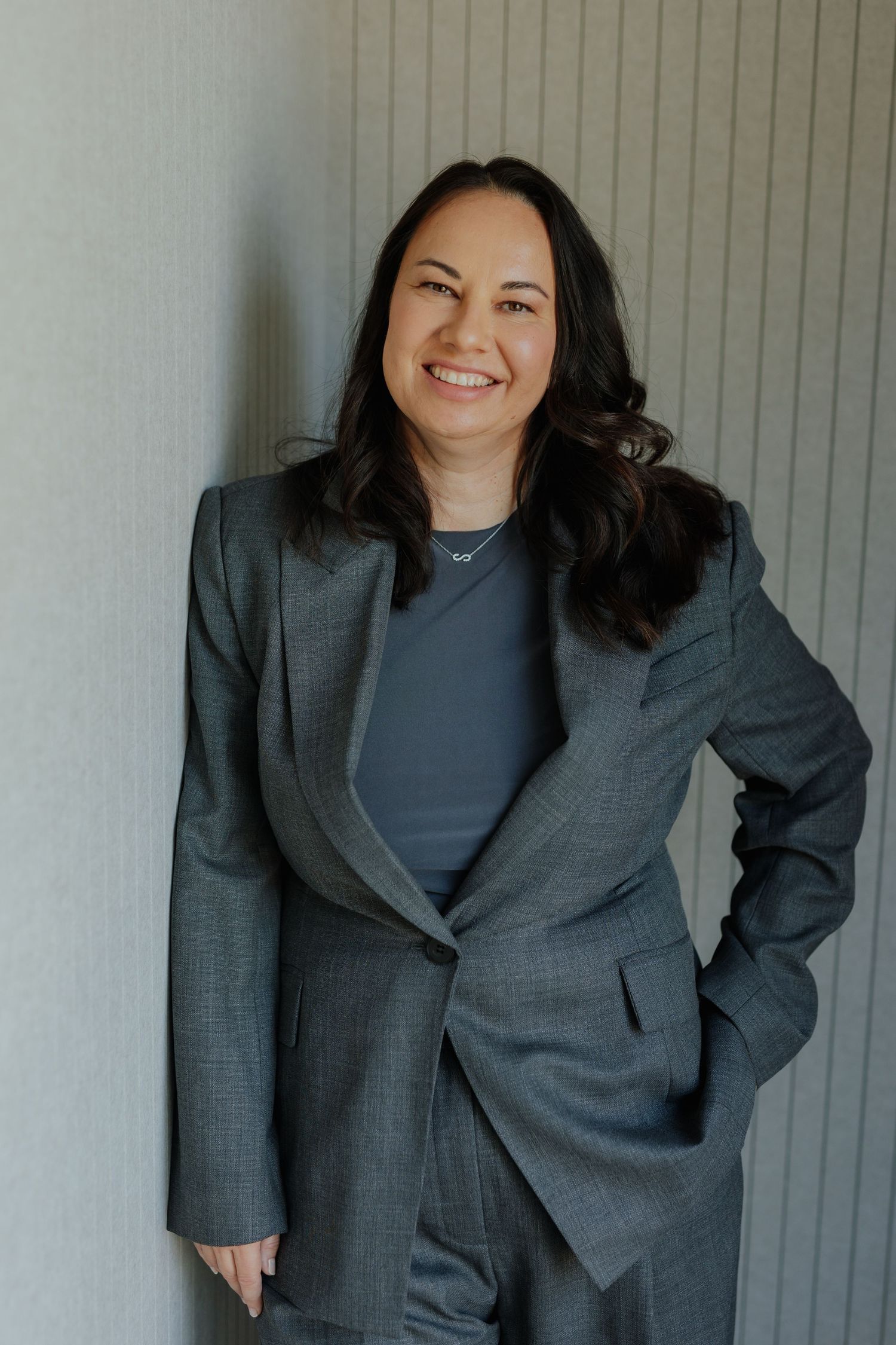 Professional woman in grey suit smiling confidently while leaning against a light textured wall.