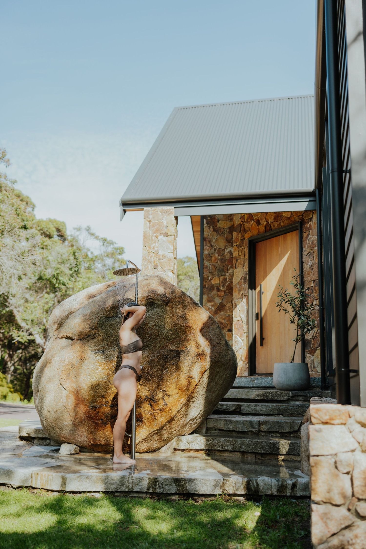 Person leaning against large boulder outside modern stone and black timber home.