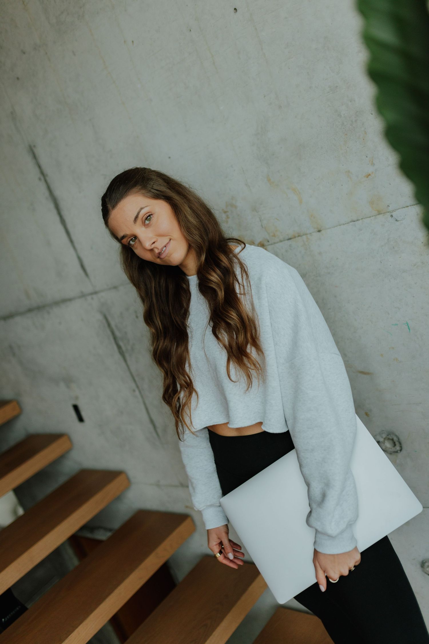 Young woman in grey crop sweatshirt holding laptop while posing on wooden stairs by concrete wall.