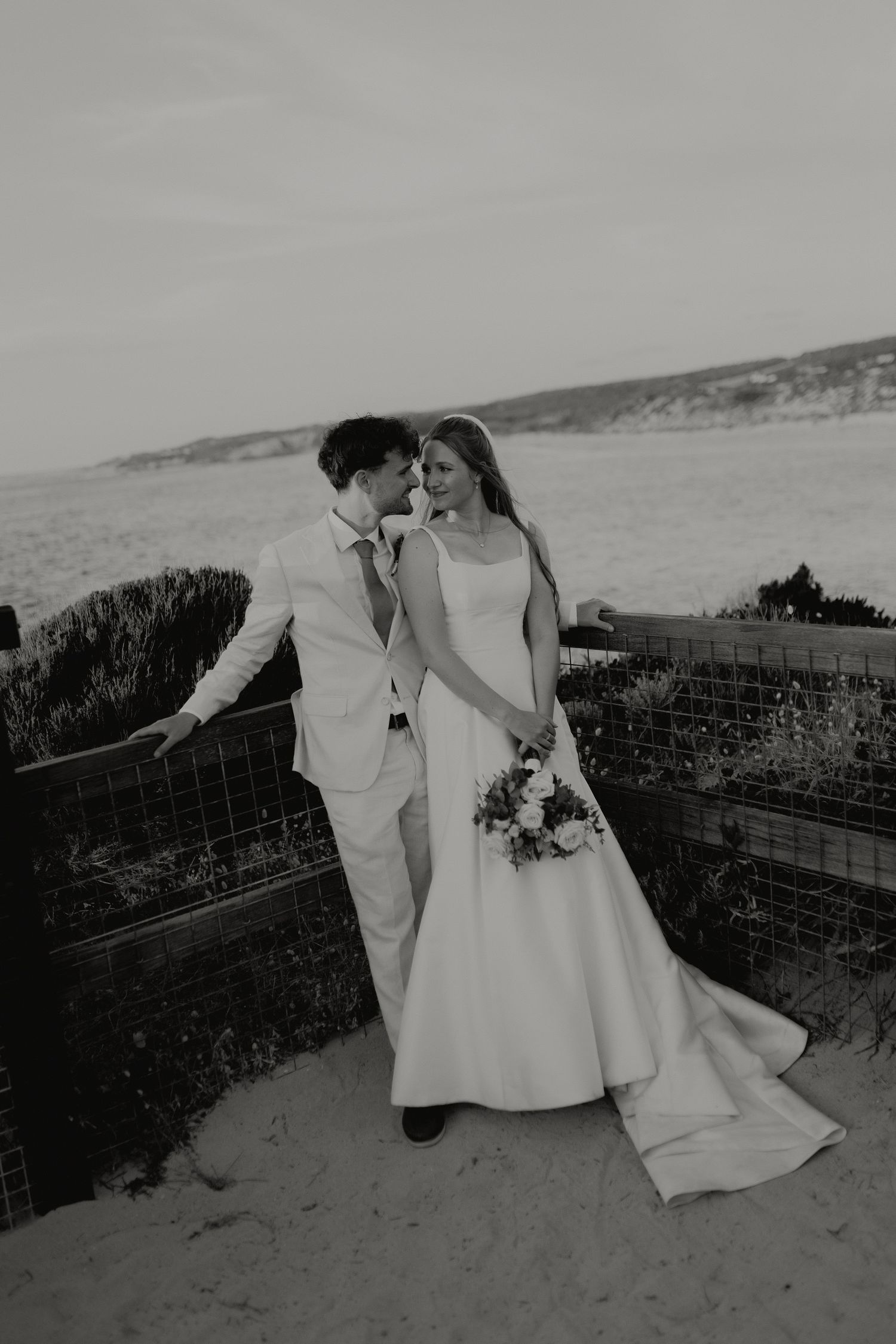 Black and white photo of bride and groom leaning close together on coastal path with ocean backdrop.