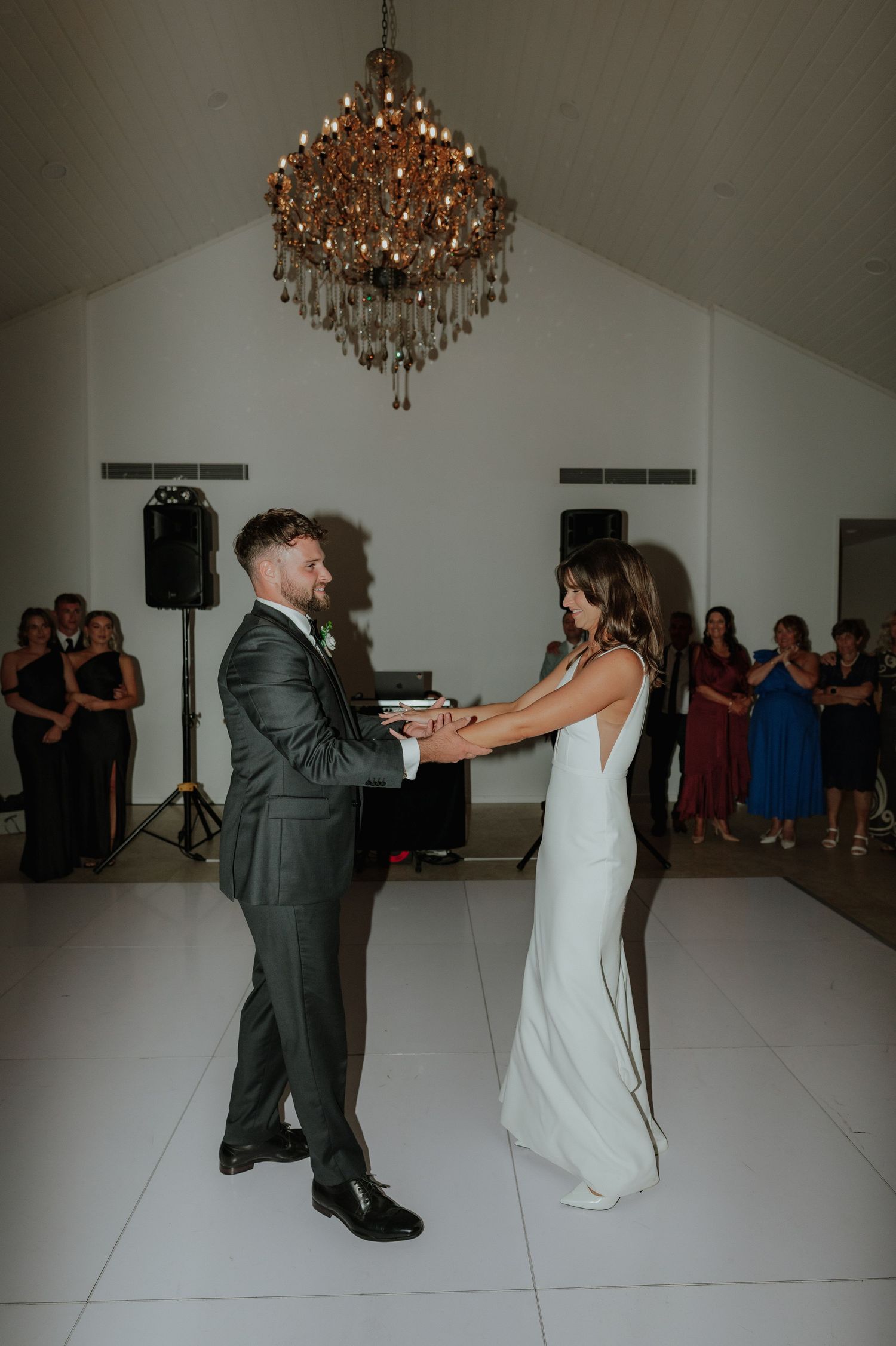 Bride and groom share first dance under ornate chandelier surrounded by wedding guests in white venue.