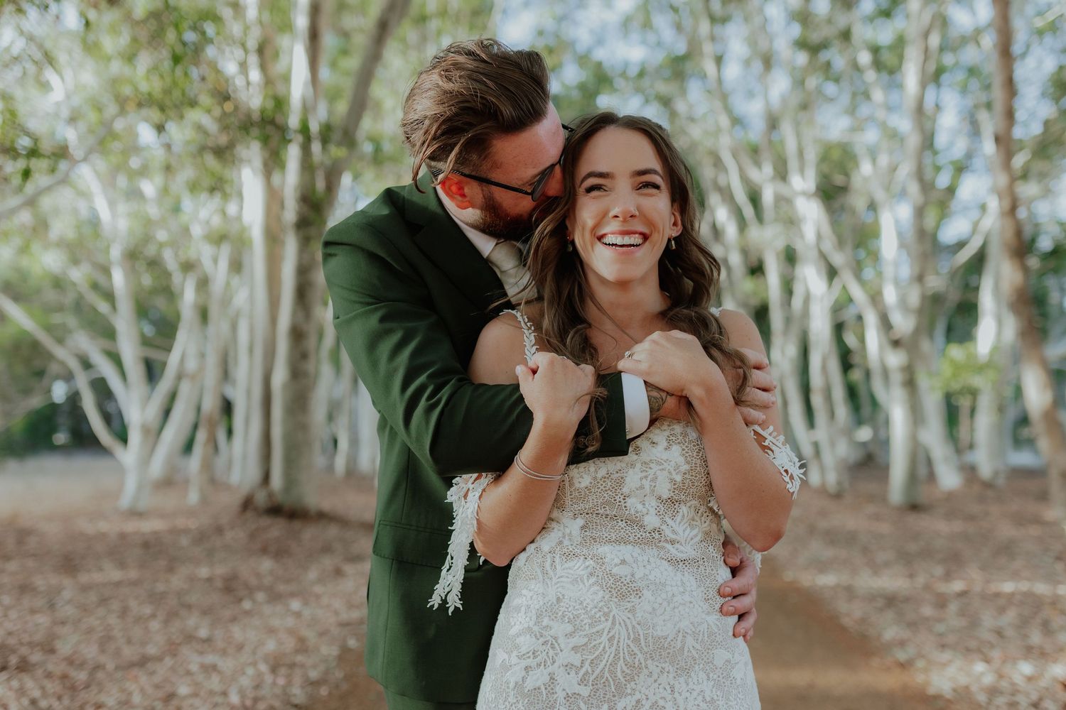 Groom in green suit embraces laughing bride in lace wedding dress among white birch trees.