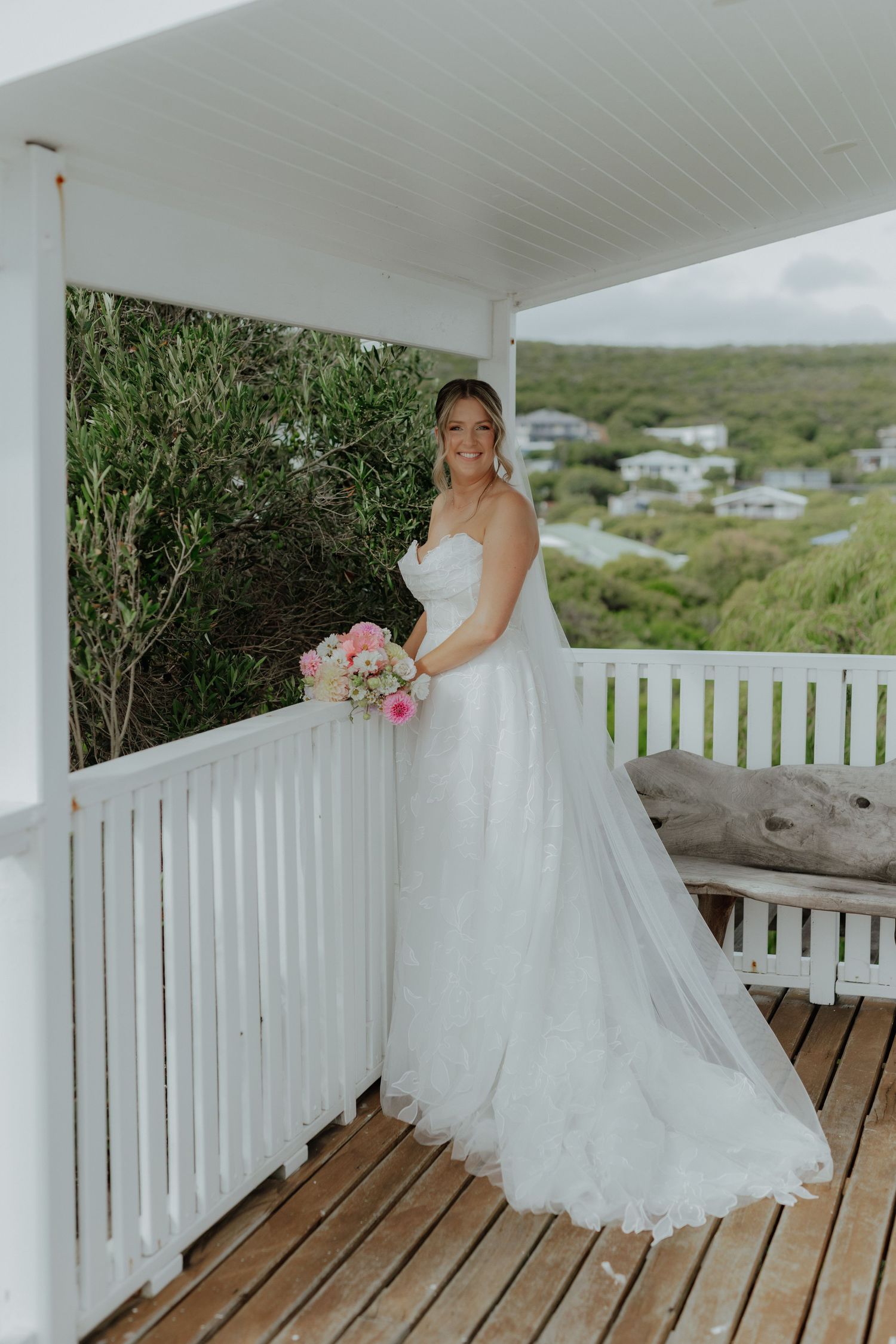 Elegant bride in strapless white tulle gown holding pink bouquet on a wooden deck balcony.