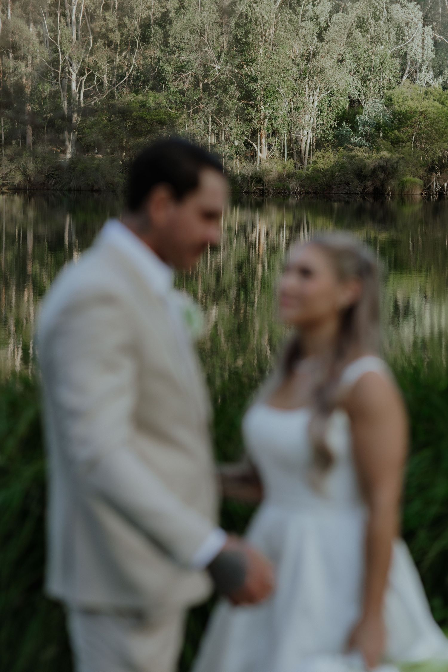Blurred artistic photo of bride and groom holding hands near a reflective lake backdrop.