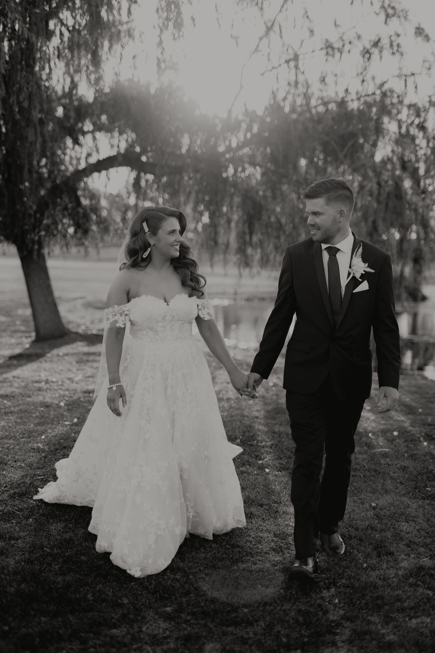 Black and white photo of bride and groom holding hands walking under weeping willow tree near water.